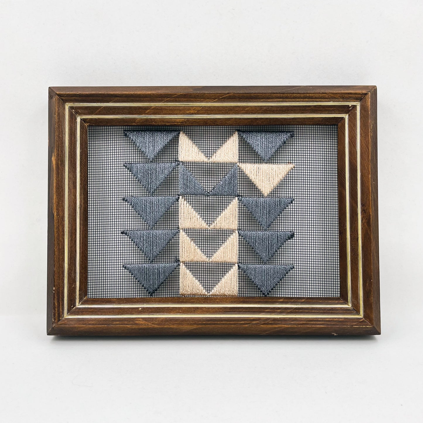 a piece of window screen hand stitched with 3 rows of triangles resembling the flying geese quilt blocks, in grey and peach, in a wood frame, on a white background