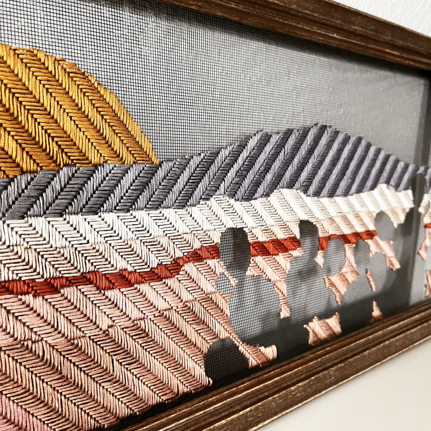 old window screen in a brown wood frame hand stitched with a landscape image of a mountain, small volcanoes and a setting sun, with prickly pears in negative space in the foreground