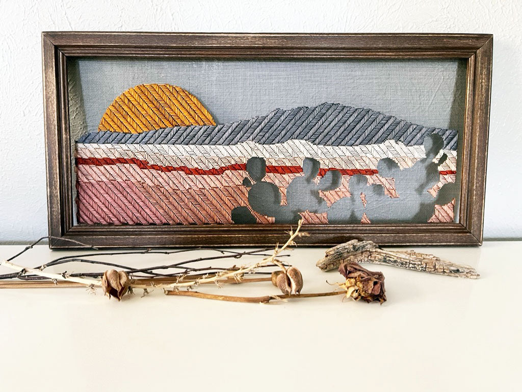old window screen in a brown wood frame hand stitched with a landscape image of a mountain, small volcanoes and a setting sun, with prickly pears in negative space in the foreground