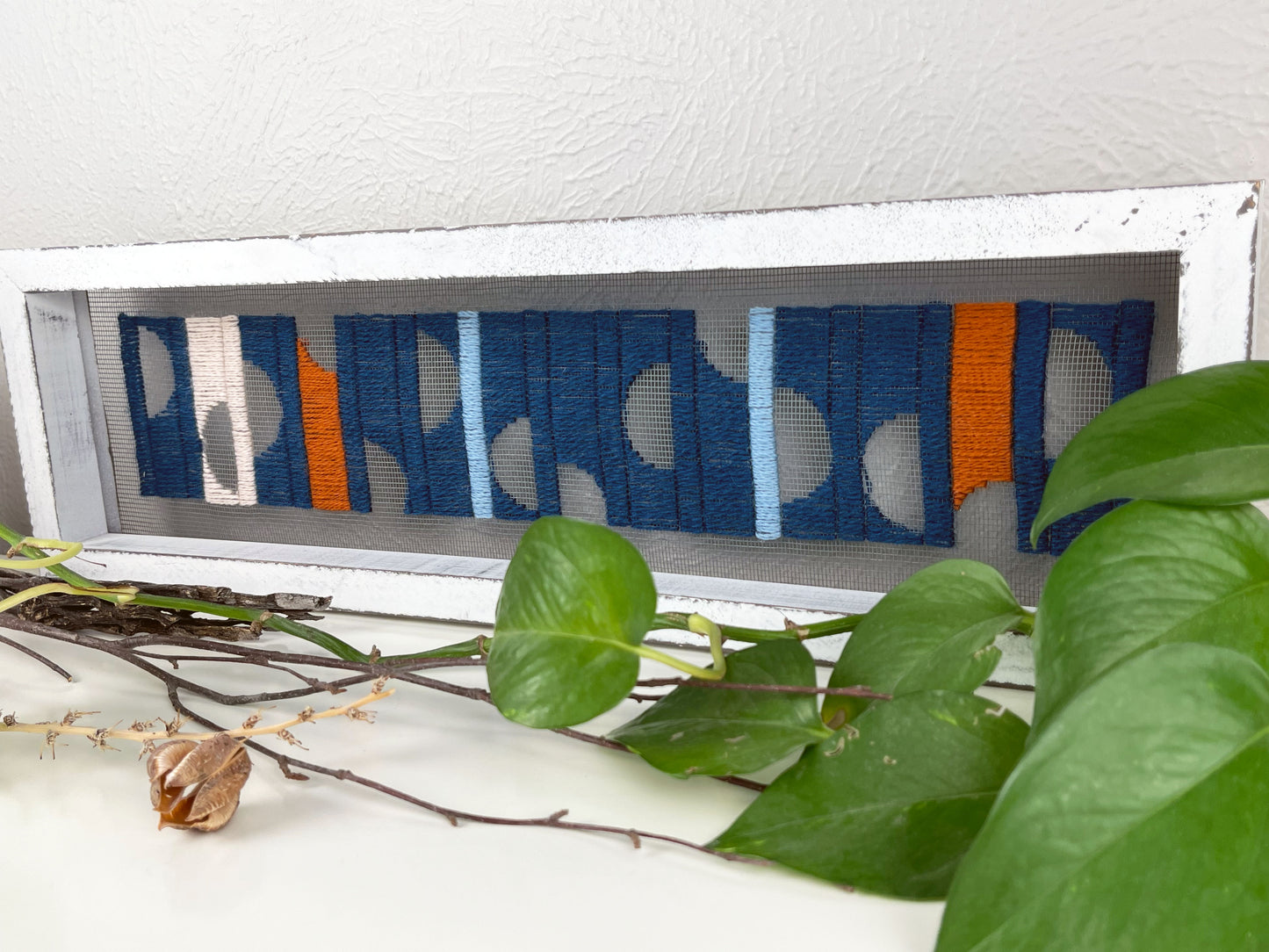 a window screen framed in a long thick white frame, the screen is hand stitched with columns of midnight blue light blue rust and peach with negative space semi circles, on a white counter with pothos vines and dried sticks around it