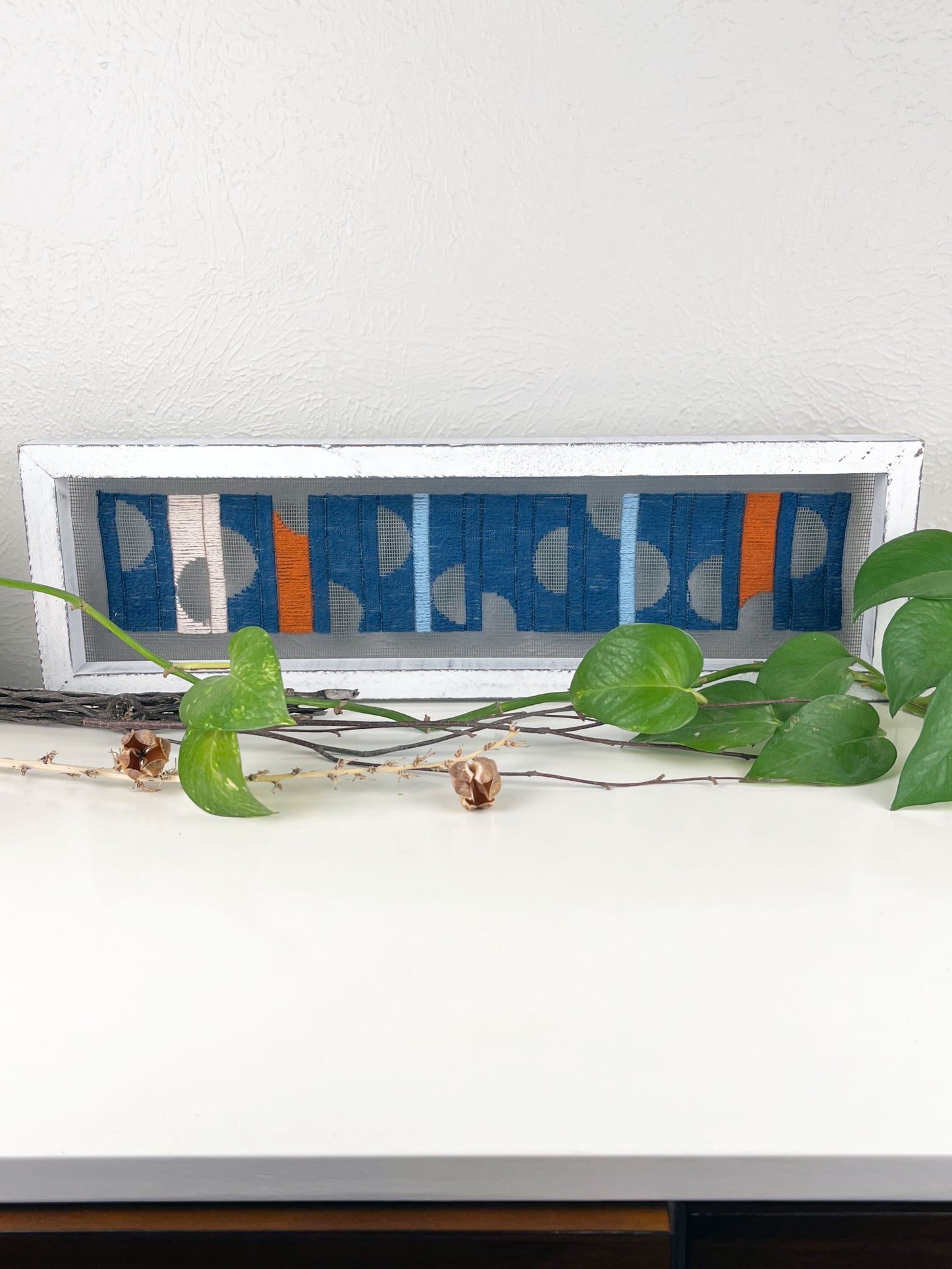 a window screen framed in a long thick white frame, the screen is hand stitched with columns of midnight blue light blue rust and peach with negative space semi circles, on a white counter with pothos vines and dried sticks around it