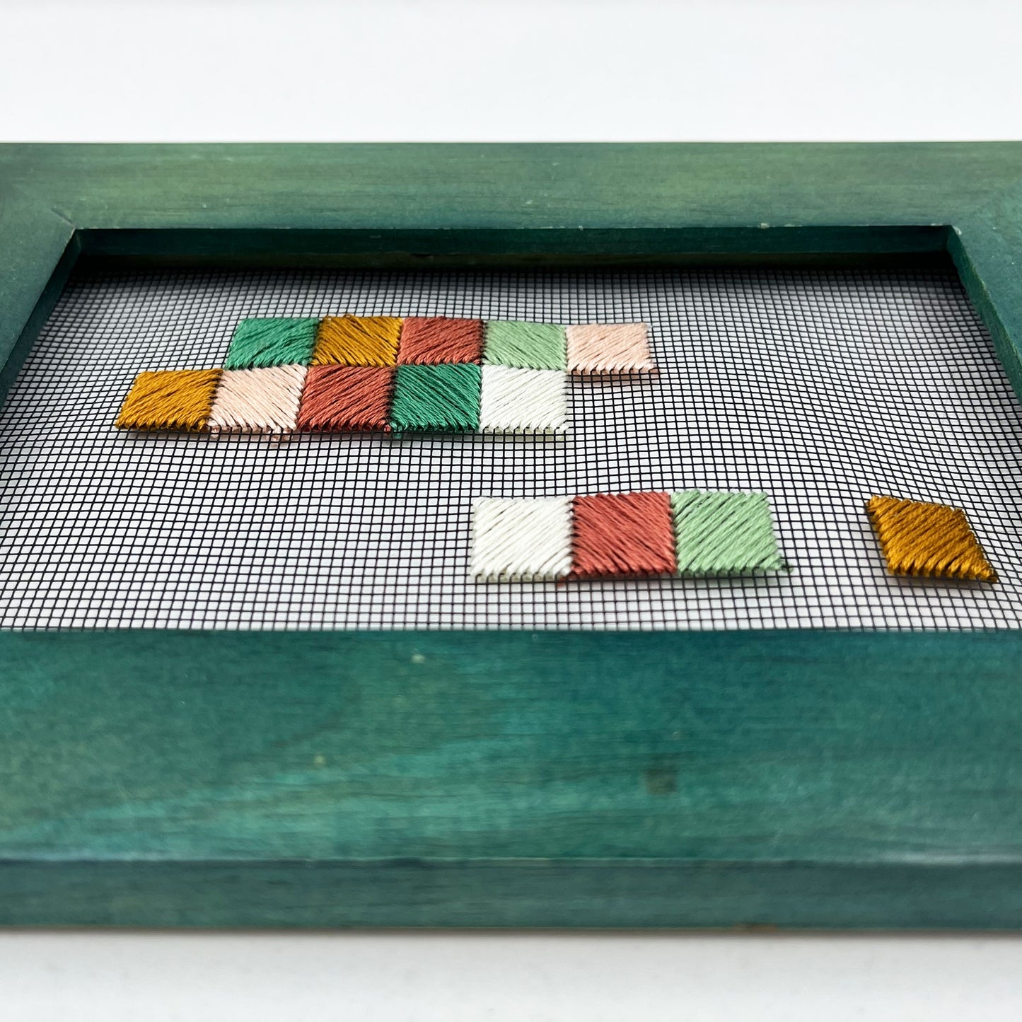 a close up angled view of a piece of window screen hand stitched with an abstract design made of squares with diagonal stitches, in greens peaches and mustard, in a wide green frame