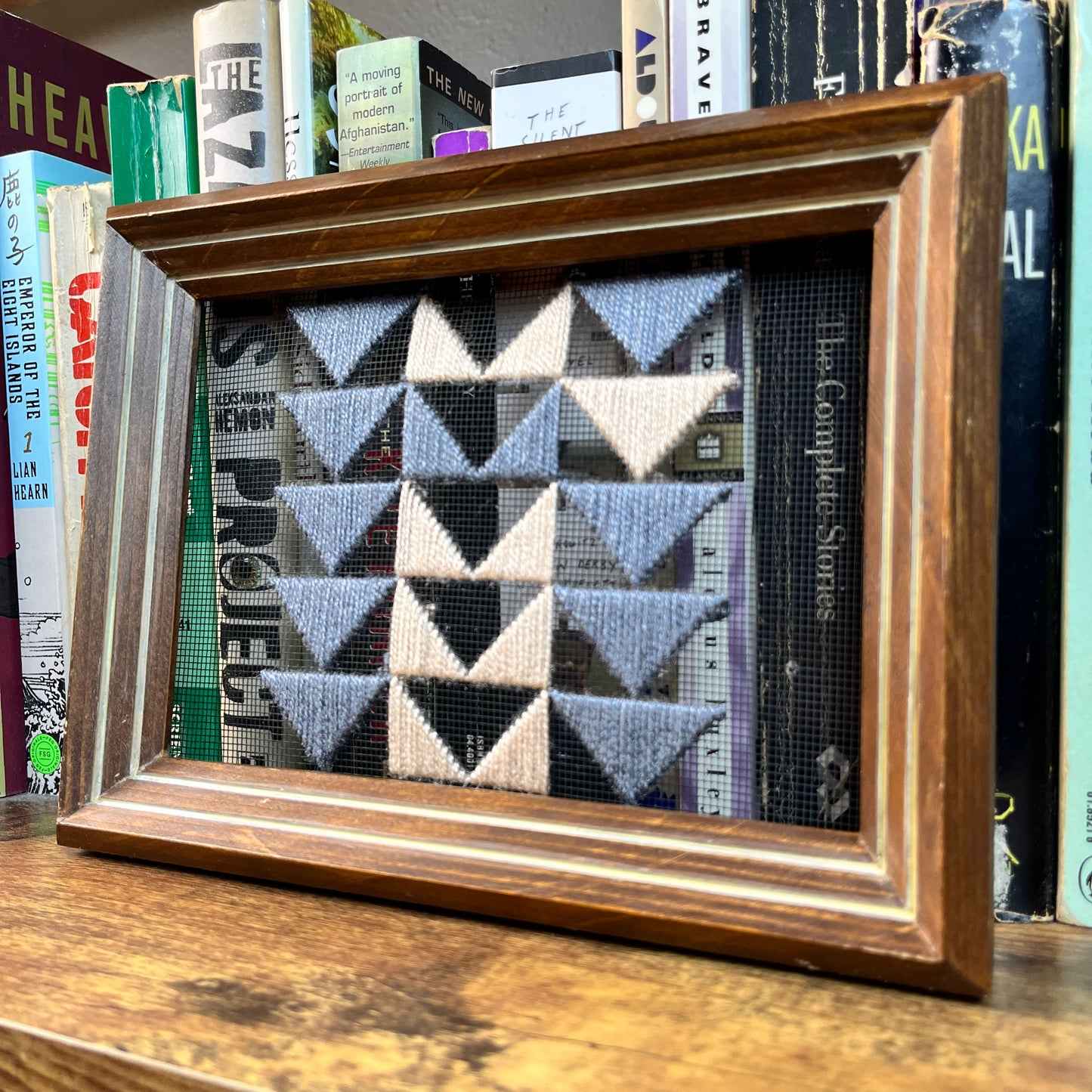 a piece of window screen hand stitched with 3 rows of triangles resembling the flying geese quilt blocks, in grey and peach, in a wood frame, perched in front of a row of books