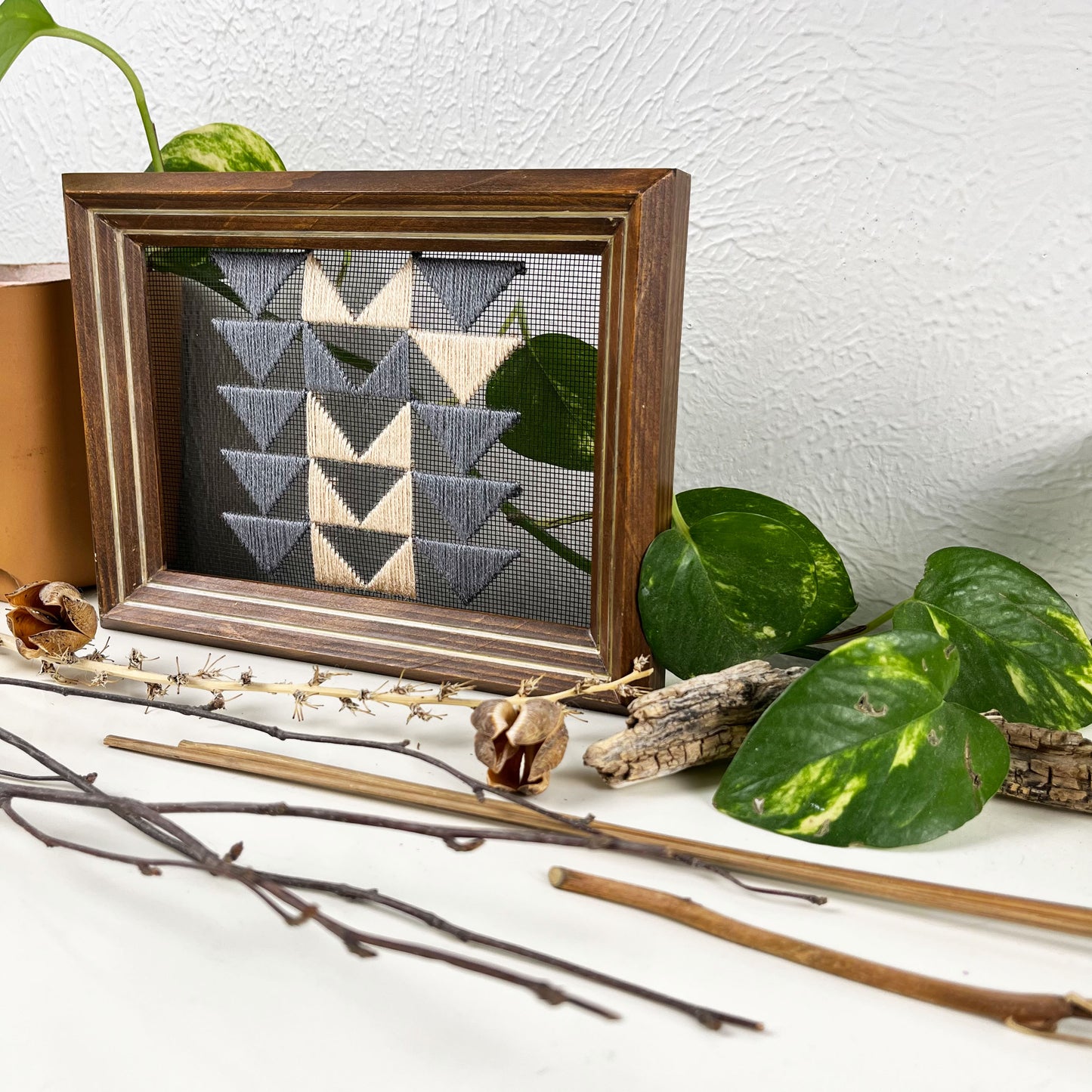 a piece of window screen hand stitched with 3 rows of triangles resembling the flying geese quilt blocks, in grey and peach, in a wood frame, on a white counter, surrounded by a pothos vine, dried flowers and sticks