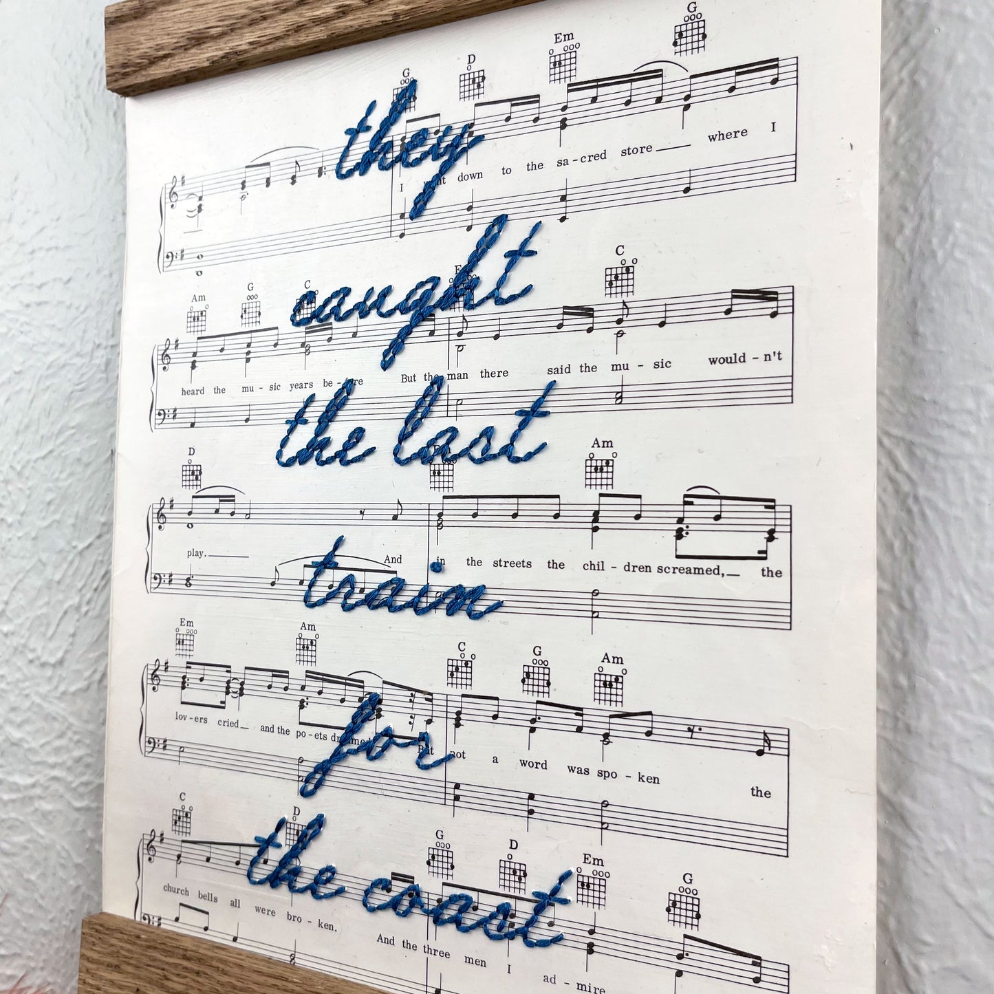 sheet music for the song American Pie by Don McLean hanging in a magnetic wood frame with the words they caught the last train for the coast hand embroidered over it in blue colored thread in cursive hanging on a wall