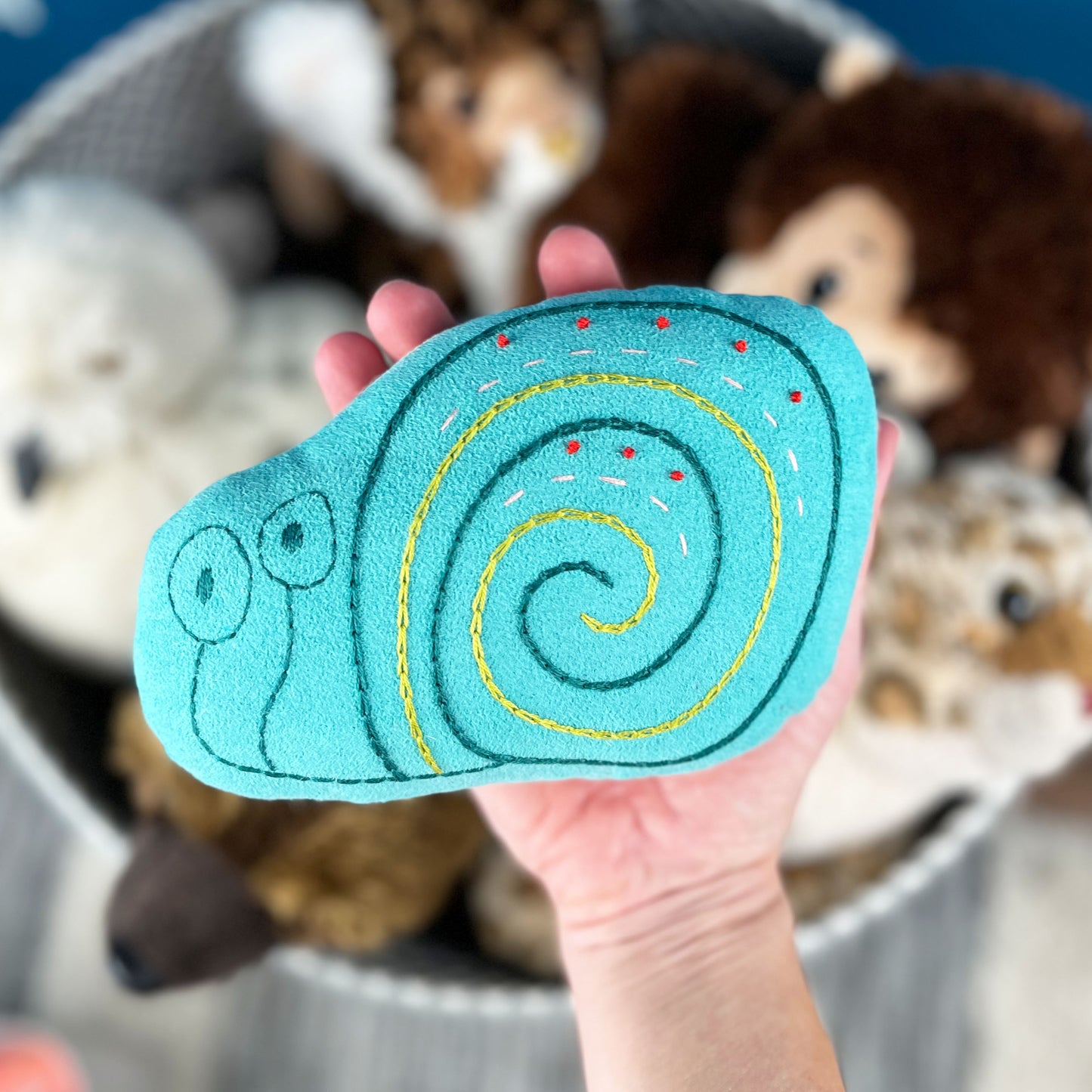a colorfully hand embroidered stuffed pillow animal of a snail in seafoam green fabric, held in a hand with out-of-focus stuffed animals in the background