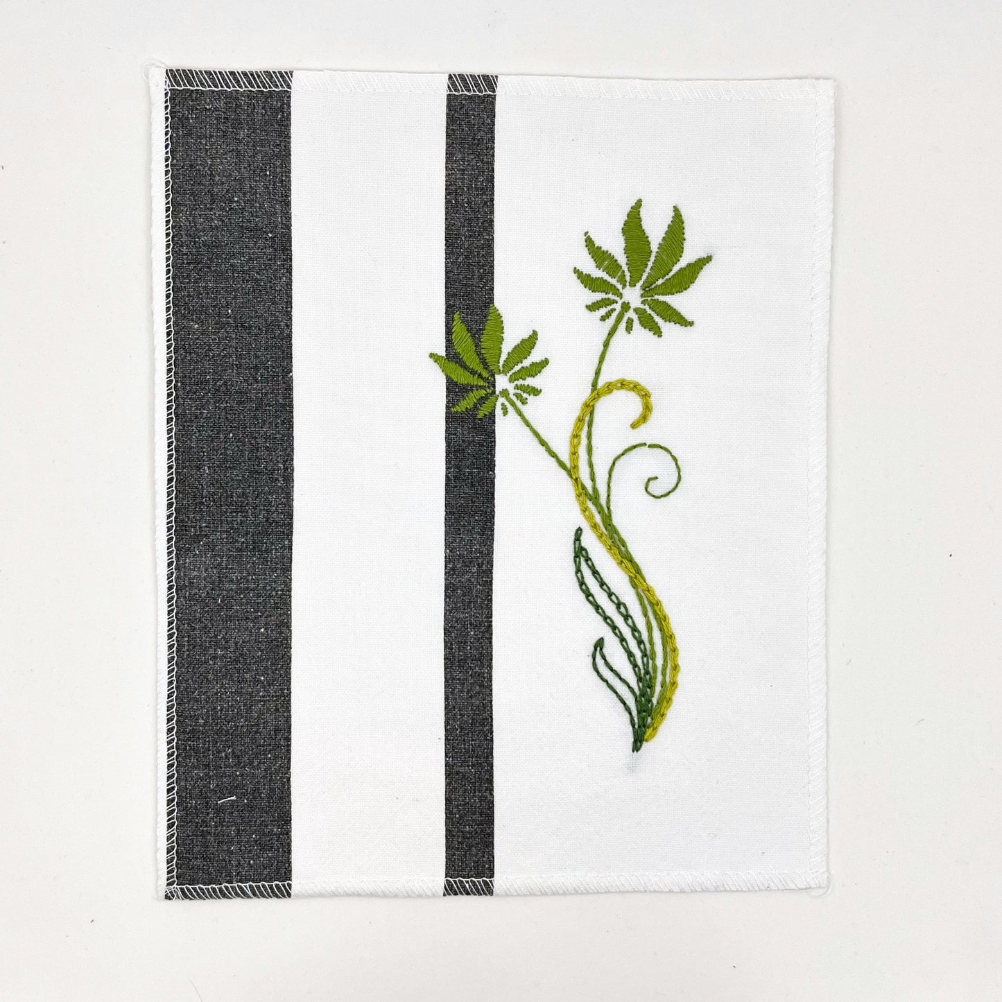 a fabric wall hanging made out of white fabric with black vertical stripes, hand stitched with flowers in shades of green with long curvy stems in chainstitch, backstitch and stem stitch, on a white background