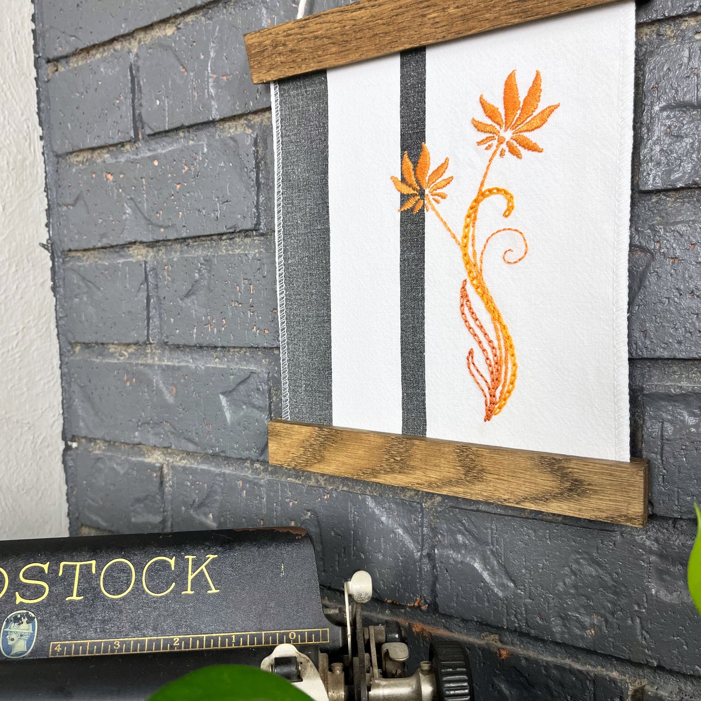 a fabric wall hanging in a wood magnetic frame on a grey wall, made out of white fabric with black vertical stripes, hand stitched with orange flowers with long curvy stems, a typewriter and pothos plant peek out at the bottom of the frame