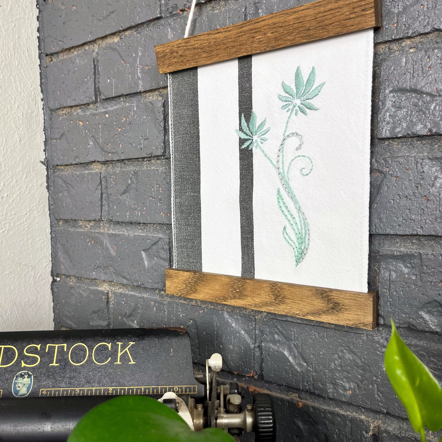 a fabric wall hanging in a wood magnetic frame on a grey wall, made out of white fabric with black vertical stripes, hand stitched with light blue flowers with long curvy stems, a typewriter and pothos plant peek out at the bottom of the frame