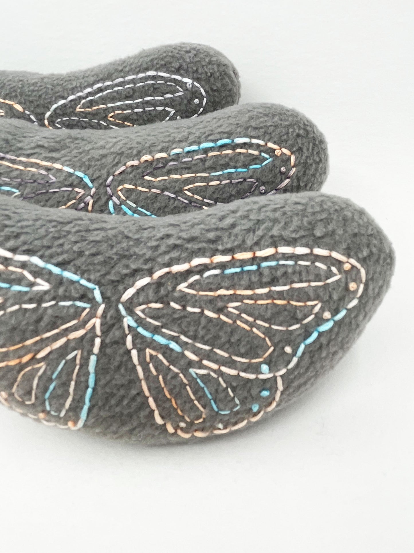 three grey fleece stuffed animals lined up from front to back, with butterfly wings hand embroidered on them in peach and sky blue variegated thread with backstitch and french knots