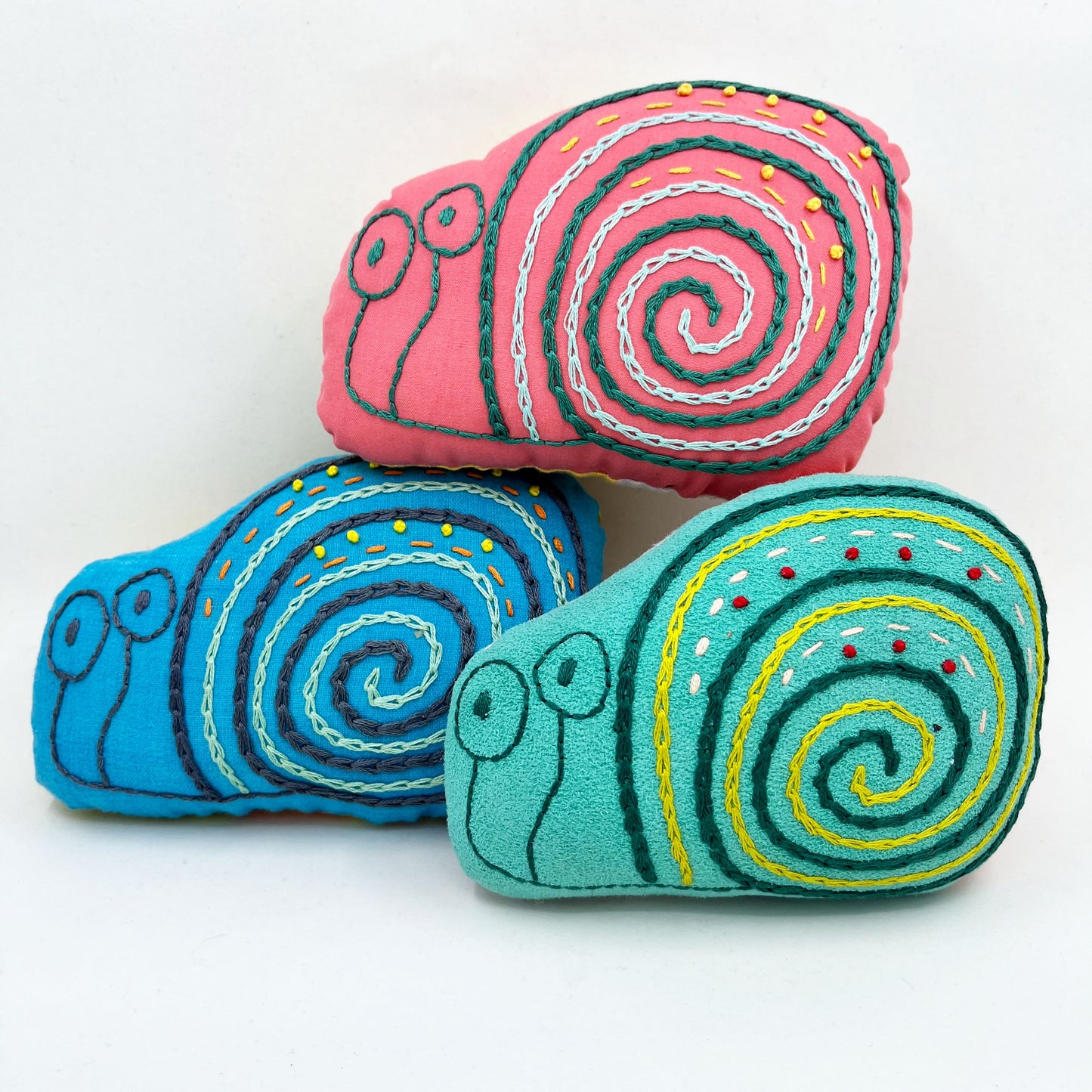 a group of colorfully hand embroidered snail stuffed animal pillows in blue, aqua and coral fabric, in a pile on a white background