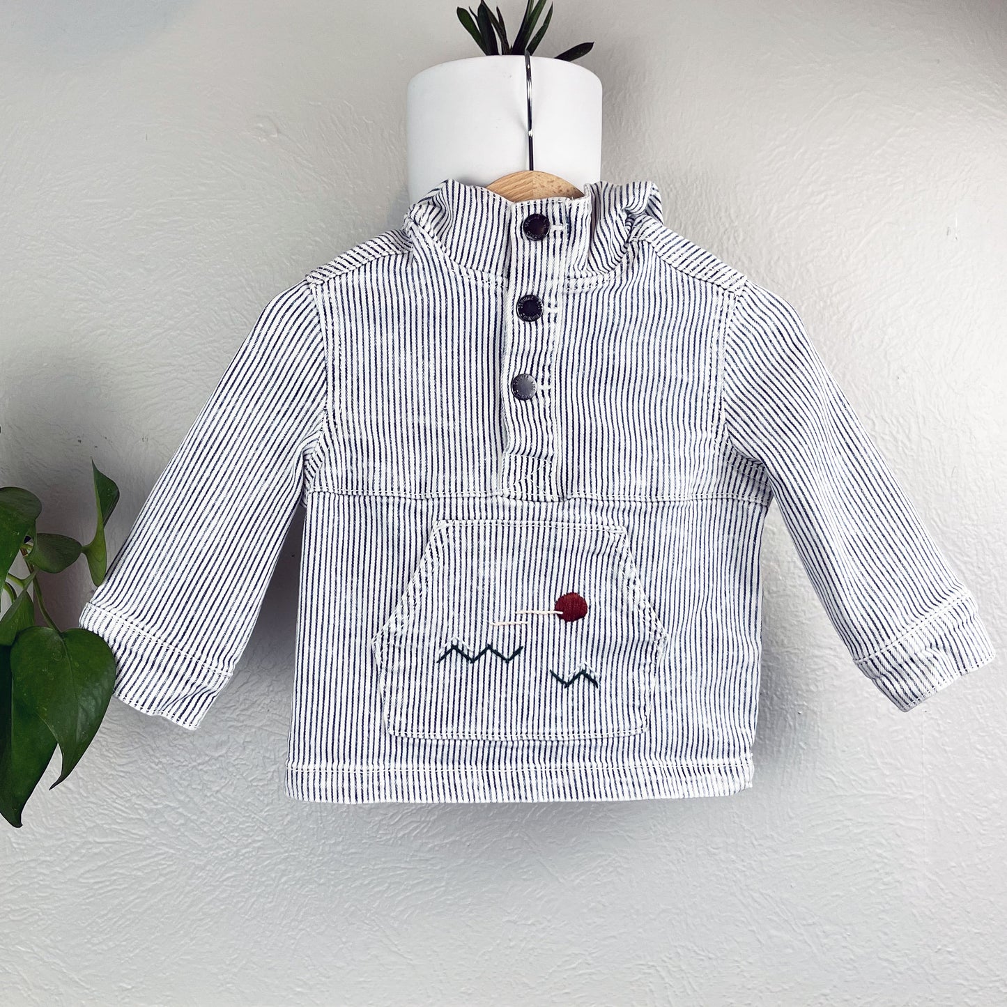 a railroad striped pullover with kangaroo pocket, with a hand embroidered abstract landscape on the pocket, hanging on a wall next to a pothos plant