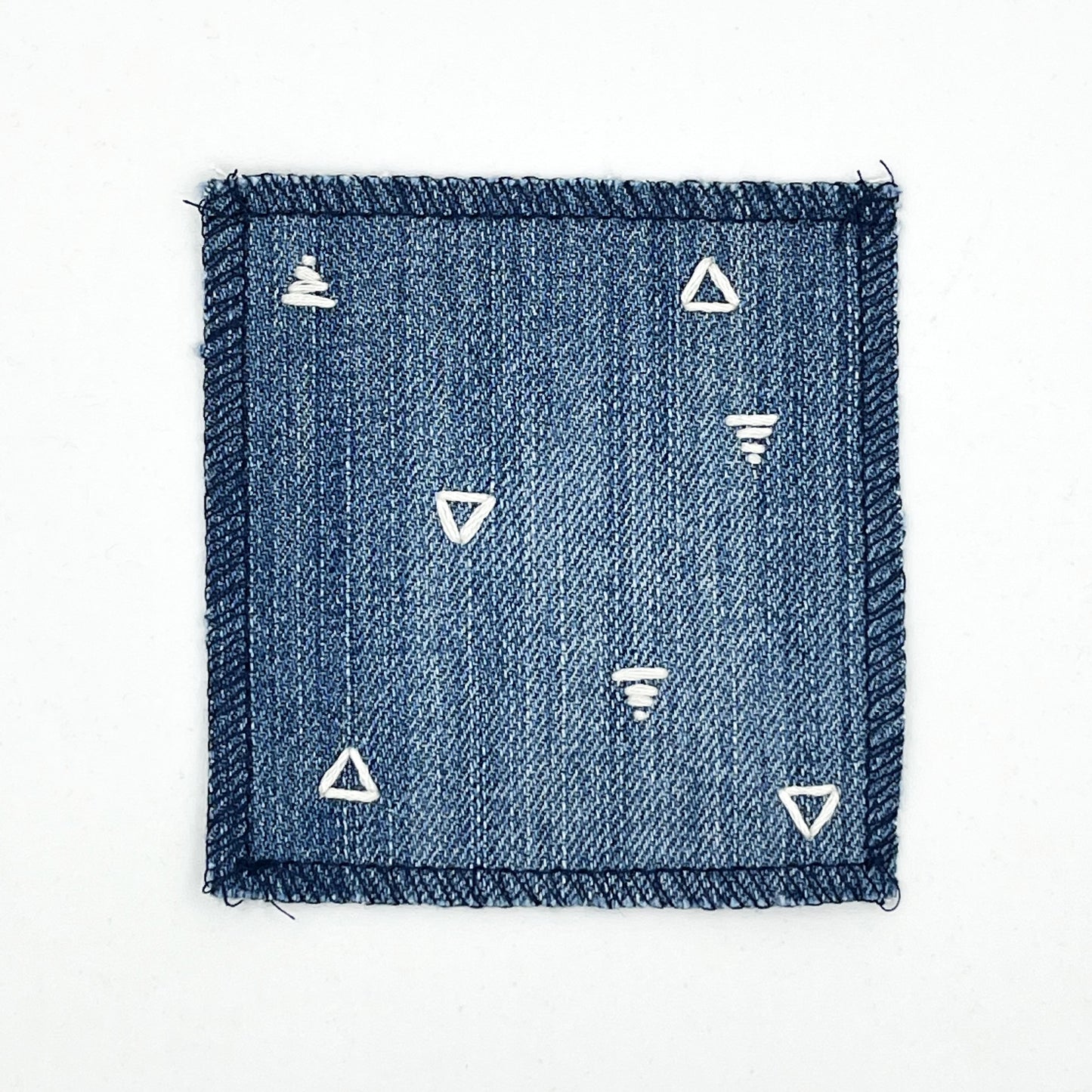 a square patch made out of denim, handstitched with scattered ivory triangles, some as outlines some with a line fill, with overlocked edges, on a white background