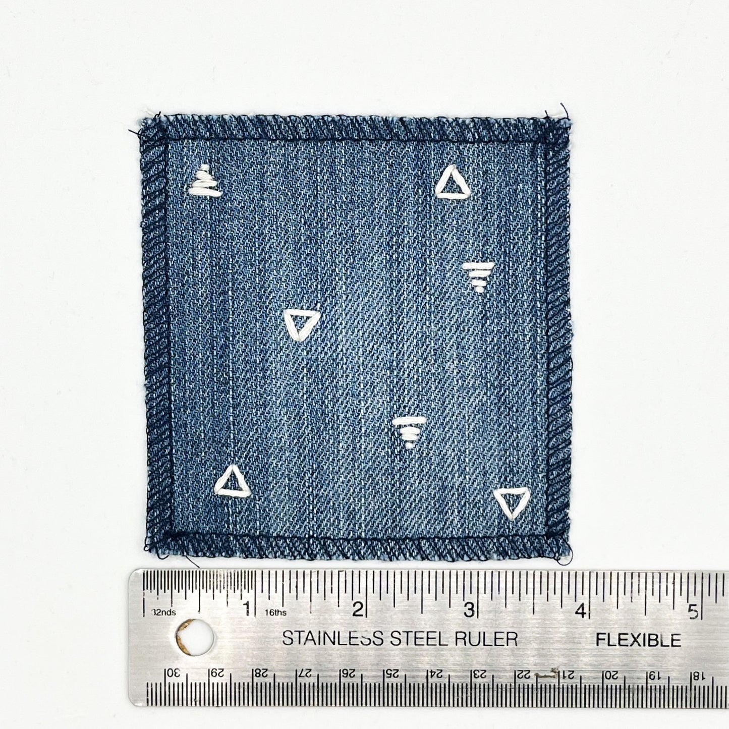 a square patch made out of denim, handstitched with scattered ivory triangles, some as outlines some with a line fill, with overlocked edges, on a white background, next to a ruler showing a width of 4 inches