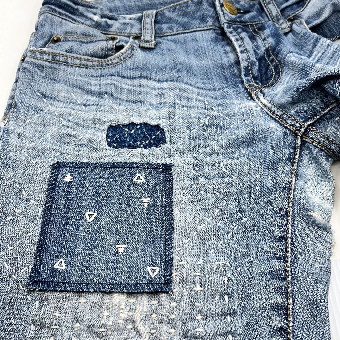 a square patch made out of denim, handstitched with scattered ivory triangles, some as outlines some with a line fill, with overlocked edges, on jeans with other visible mending and sashiko stitching