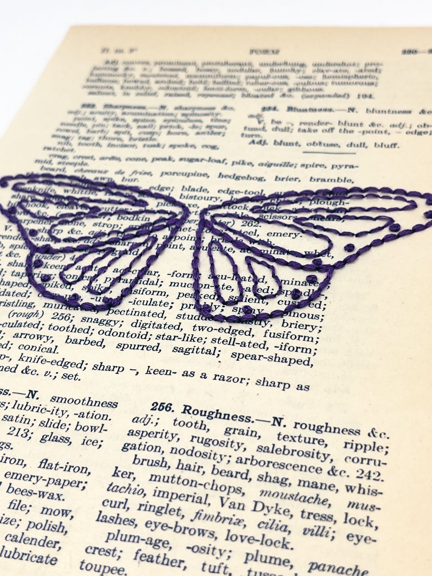 close up view of a vintage thesaurus page hand embroidered in eggplant colored thread with butterfly wings, on a white background
