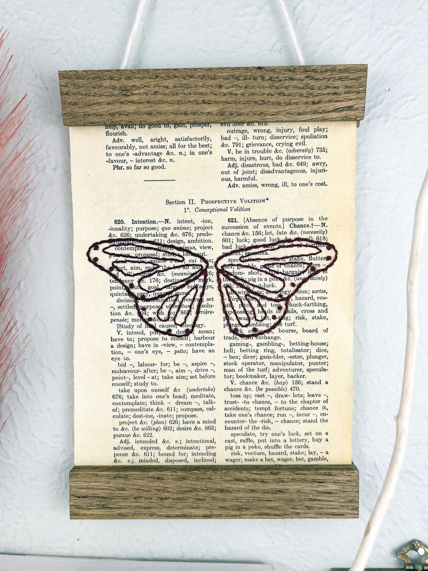 a vintage thesaurus page in a magnetic wood frame hanging on a wall, the book page is hand embroidered in plum colored thread with butterfly wings