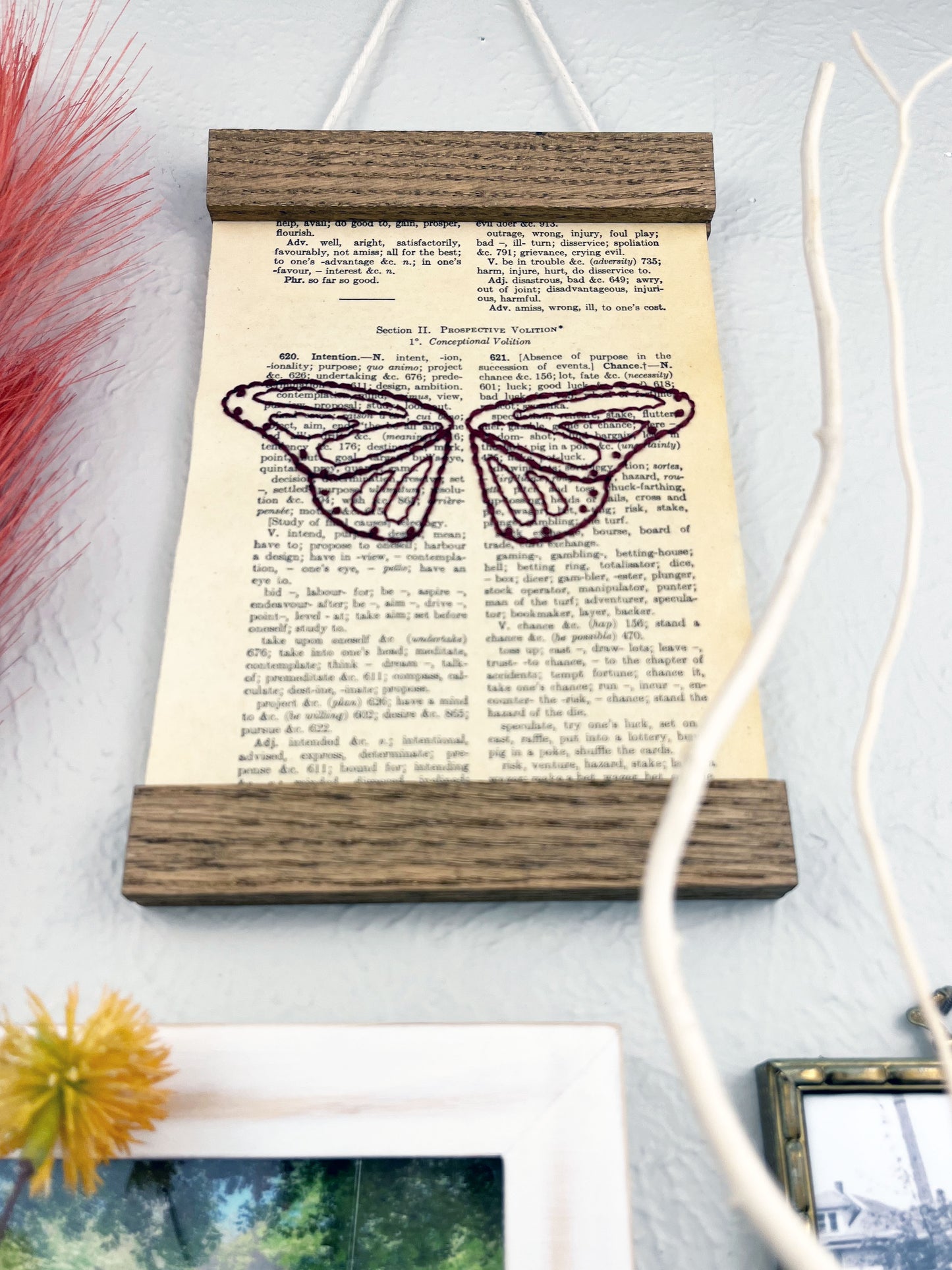 low angle view of a vintage thesaurus page in a magnetic wood frame hanging on a wall next to other framed photos and decorative branches and feathers, the book page is hand embroidered in plum colored thread with butterfly wings