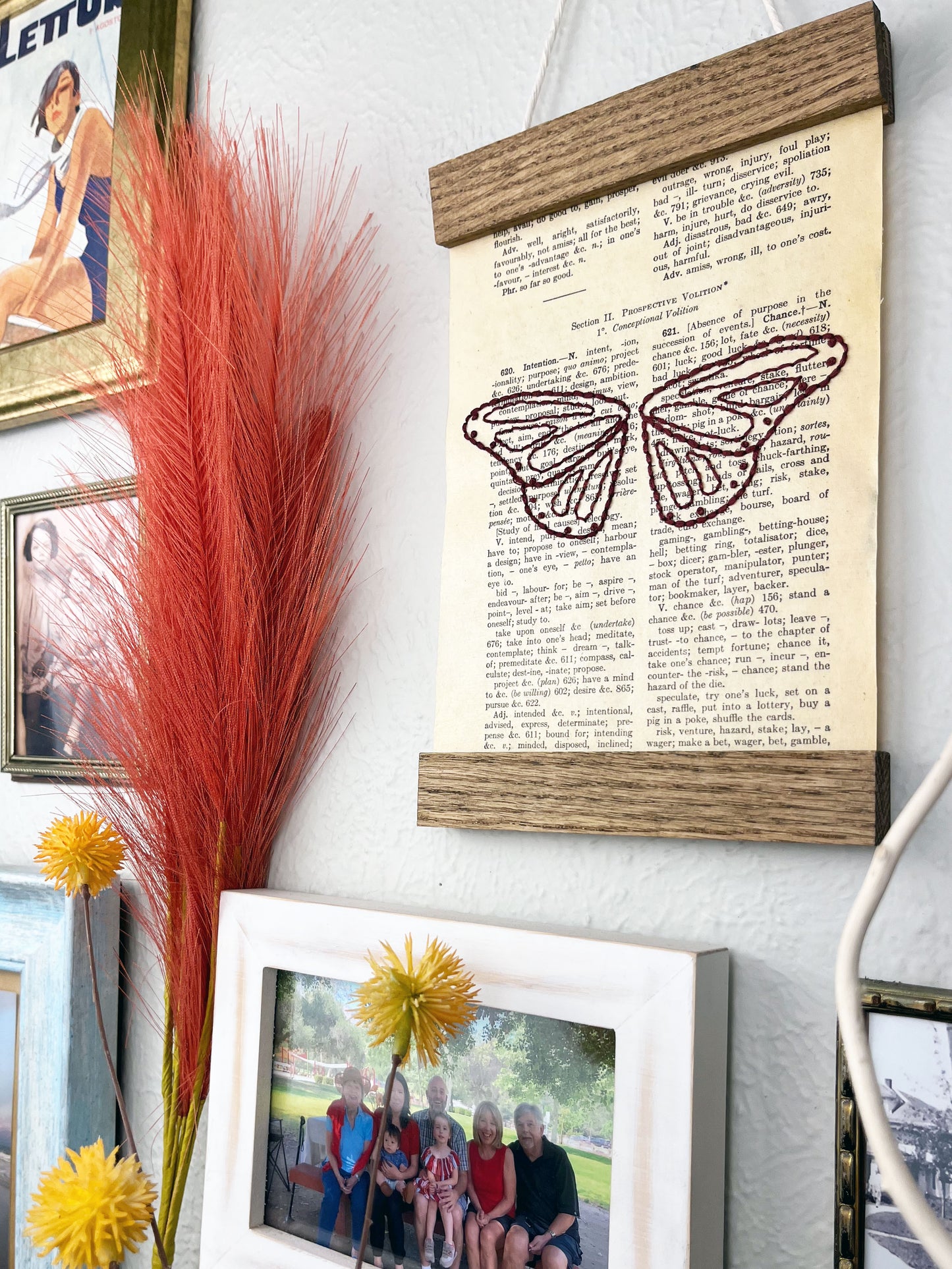 a vintage thesaurus page in a magnetic wood frame hanging on a wall next to other framed photos and decorative branches and feathers, the book page is hand embroidered in plum colored thread with butterfly wings