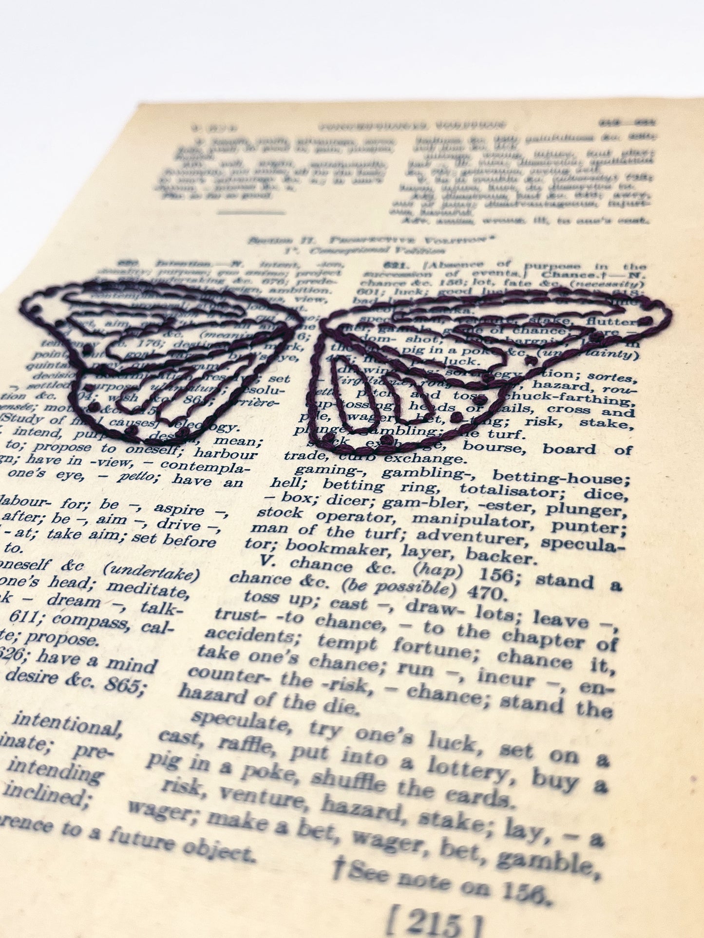 close up angled view of a vintage thesaurus page hand embroidered in eggplant colored thread with butterfly wings, on a white background