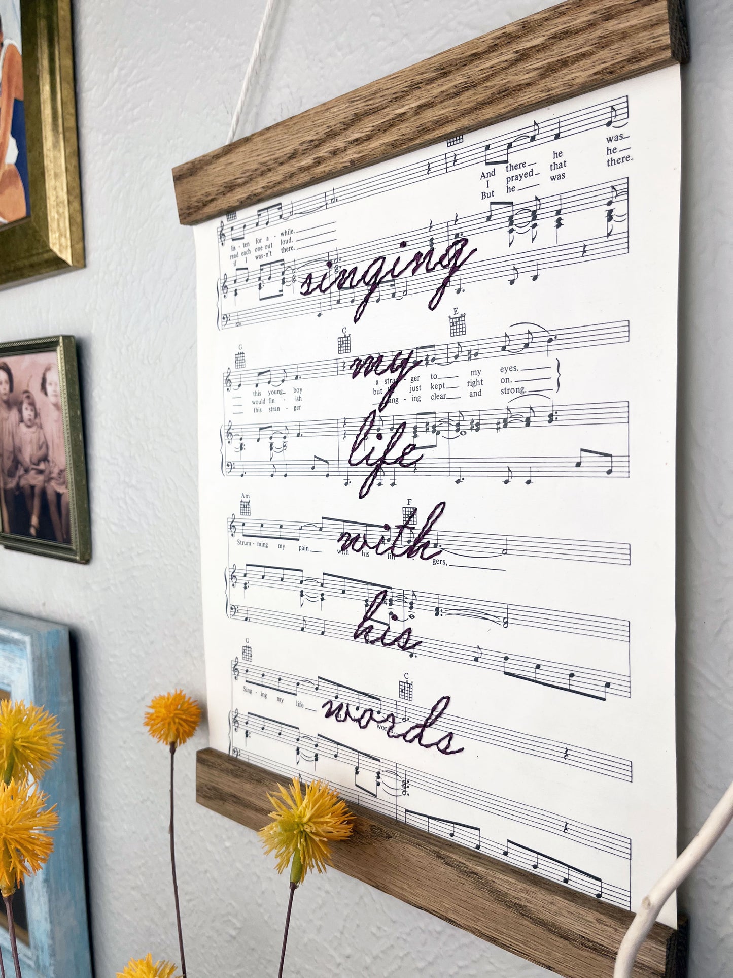 angled view of sheet music for the song Killing Me Softly With His Words hanging in a magnetic wood frame with the words singing my life with his words hand embroidered over it in eggplant colored thread in cursive, hanging on a wall with other framed photos and decorative flowers and twigs in front of it