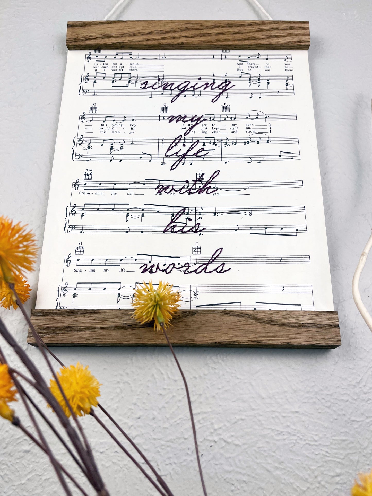 low angle view of sheet music for the song Killing Me Softly With His Words hanging in a magnetic wood frame with the words singing my life with his words hand embroidered over it in eggplant colored thread in cursive, hanging on a wall with decorative flowers and twigs in front of it