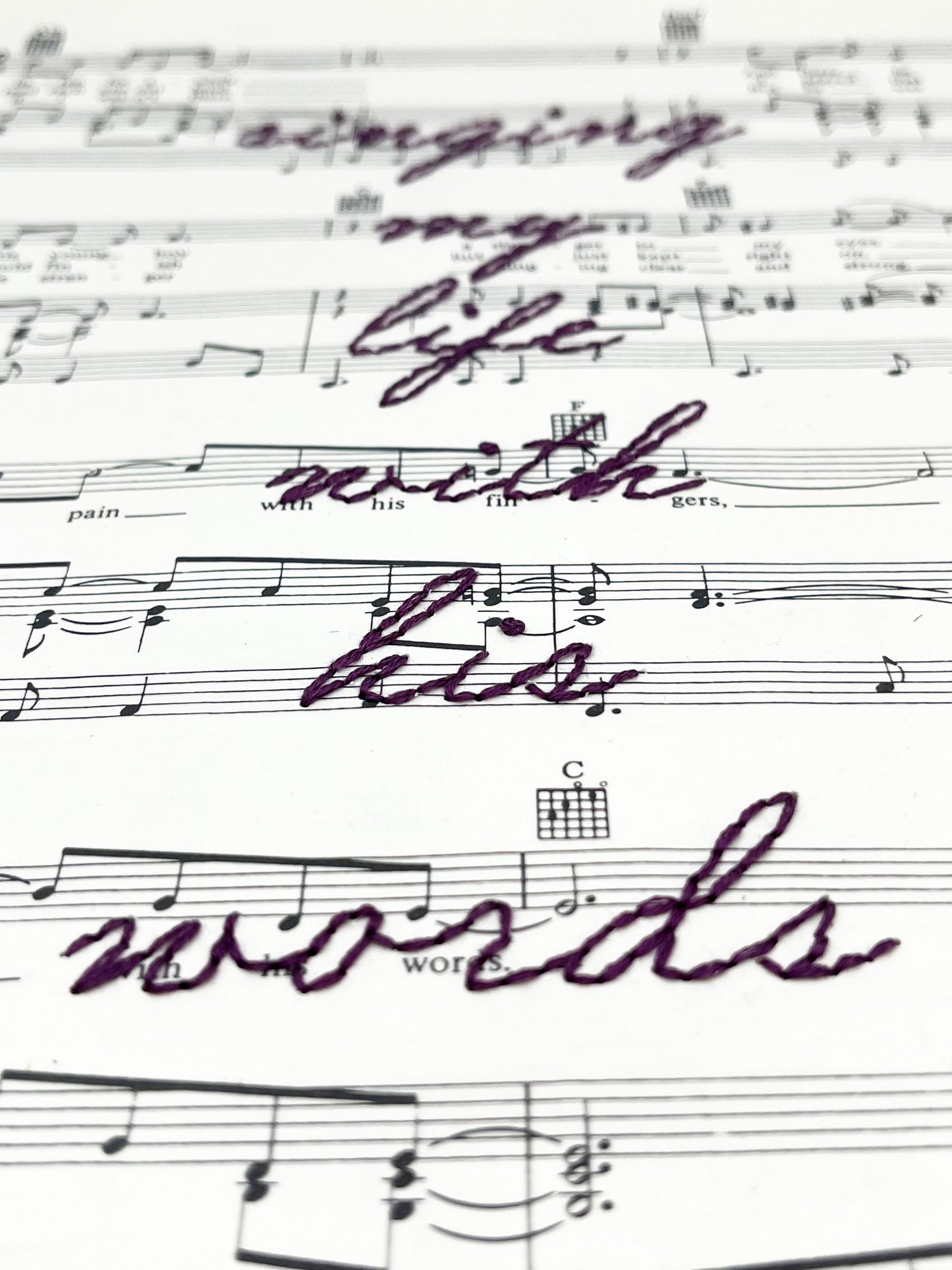 close up view of sheet music for the song Killing Me Softly With His Words with the words singing my life with his words hand embroidered over it in eggplant colored thread in cursive, on a white background