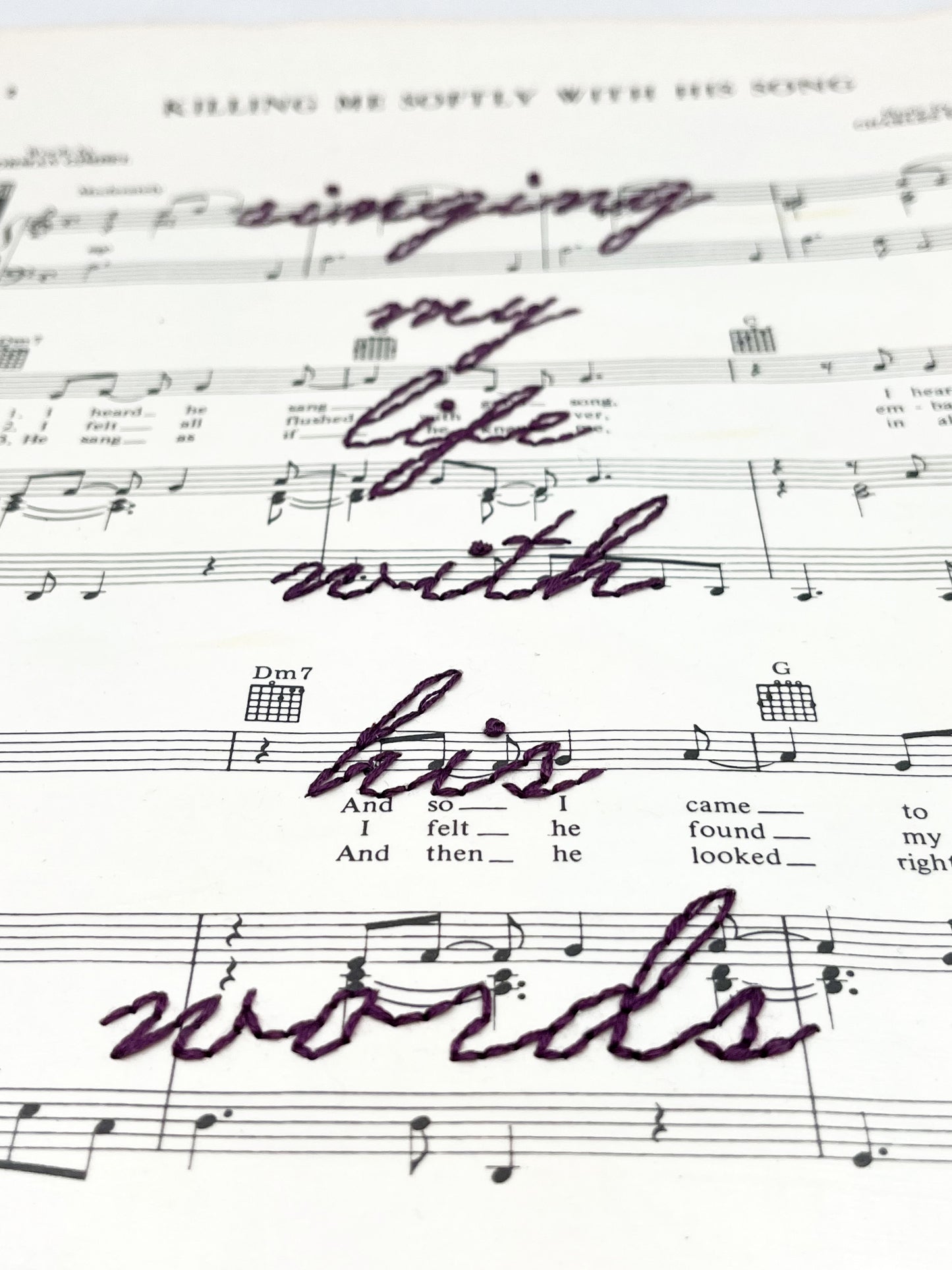 close up view of sheet music for the song Killing Me Softly With His Words with the words singing my life with his words hand embroidered over it in eggplant colored thread in cursive, on a white background