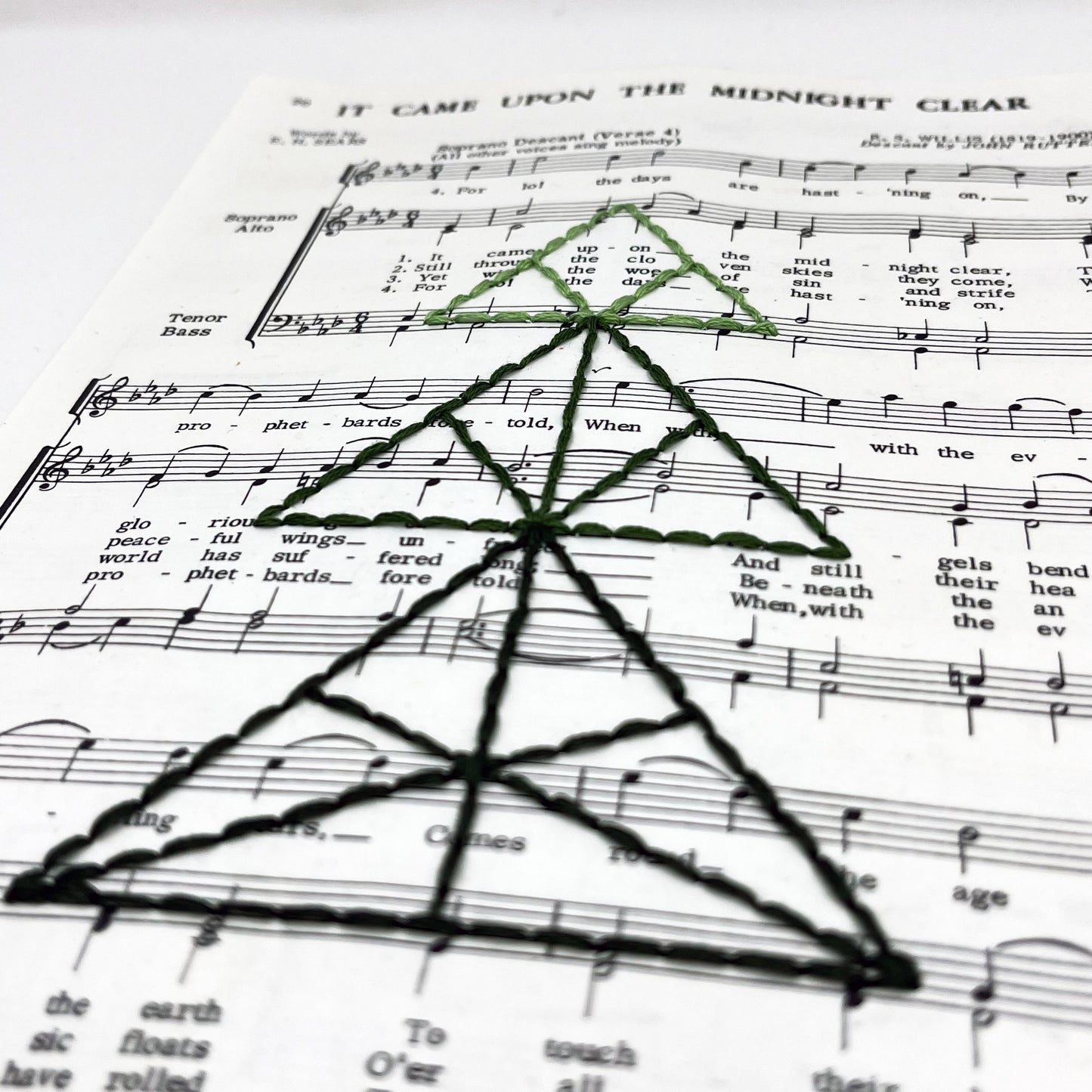 a close up angled view of sheet music from the Christmas song "It Came Upon the Midnight Clear", hand stitched over with a Christmas tree made from triangles, in shades of green thread
