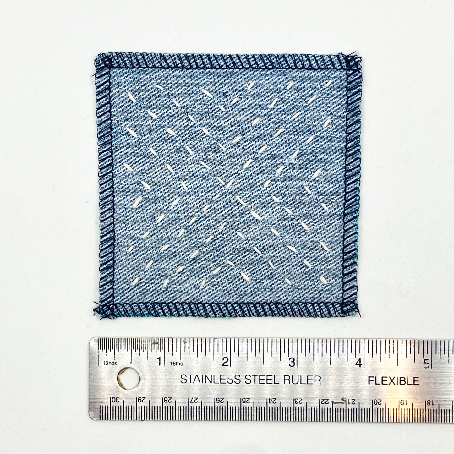 a square patch made out of denim, handstitched with ivory running stitches in a radiating X pattern, with overlocked edges, next to a metal ruler to show a width of 4 inches, on a white background