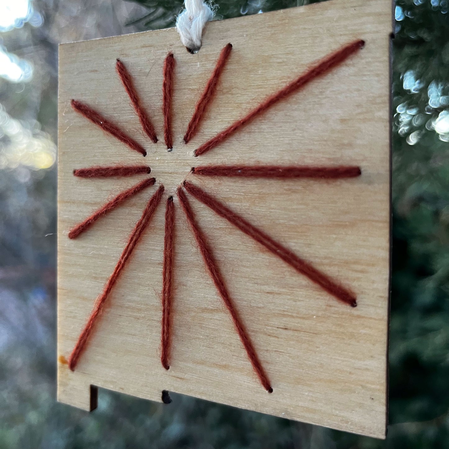 close up of a laser cut wood ornament in the shape of New Mexico, hand embroidered in red with negative space in the shape of a heart where Albuquerque is, and lines coming out from it like sunbeams, with an off white cord loop hanging on a pine tree