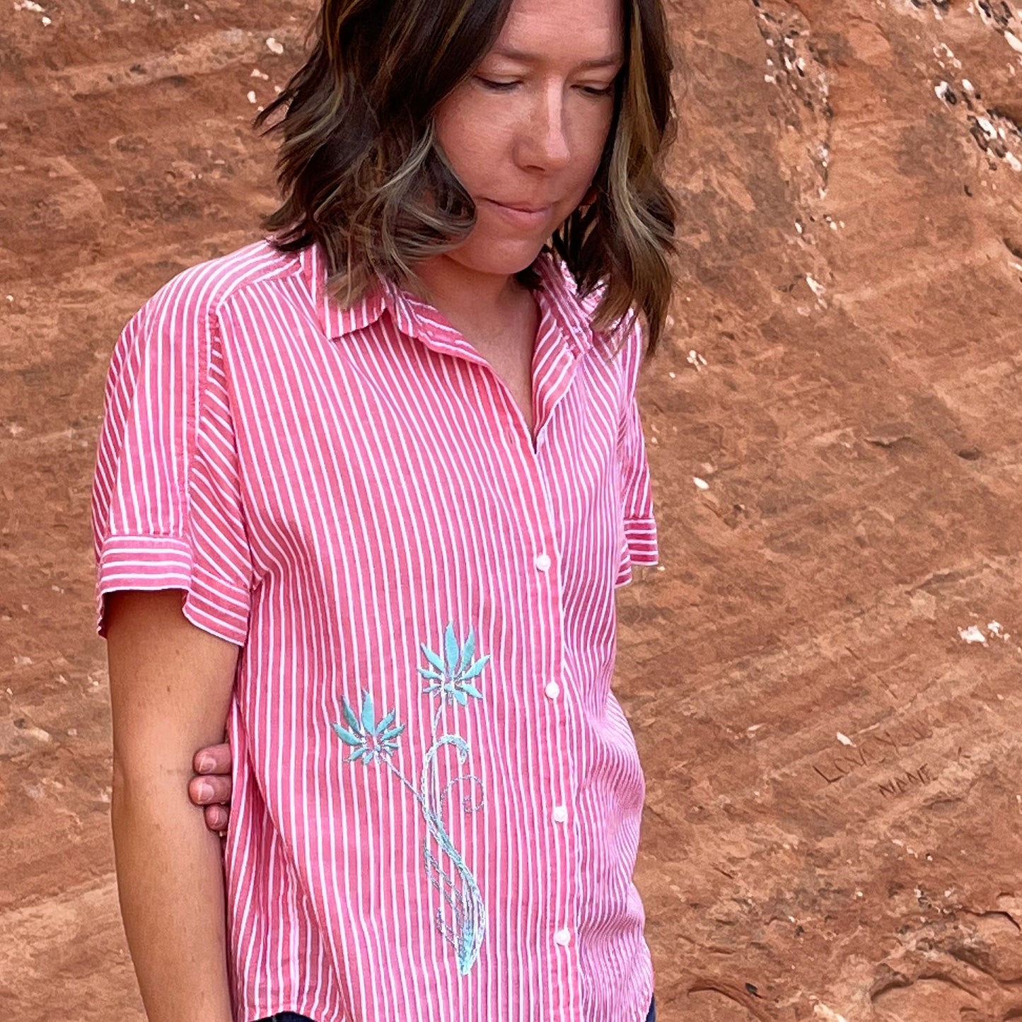 a woman from the waist up in a red and white striped button-up short sleeve shirt, with two hand embroidered long stemmed flowers in aqua on the lower front right side of the shirt