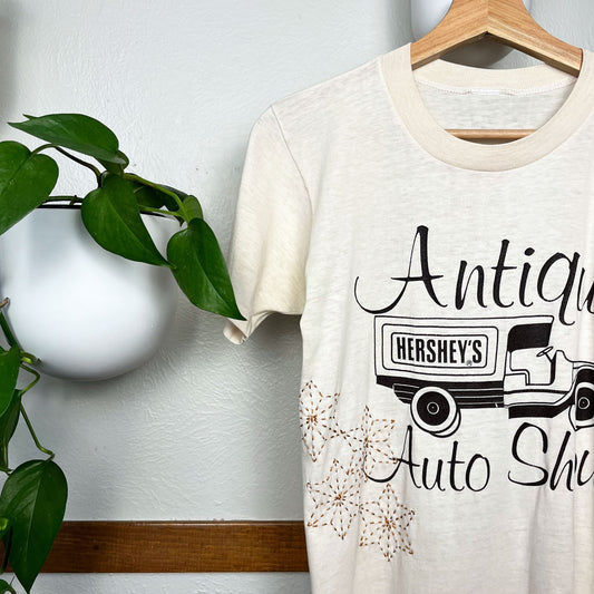 Cream colored T-shirt hanging on a hanger next to a pothos plant on a wall, the t-shirt has a graphic print of a truck that says Hershey's on the back, and the words Antique Auto Show printed around it, on the bottom side of the tee there are hand embroidered sashiko stars in mustard brown