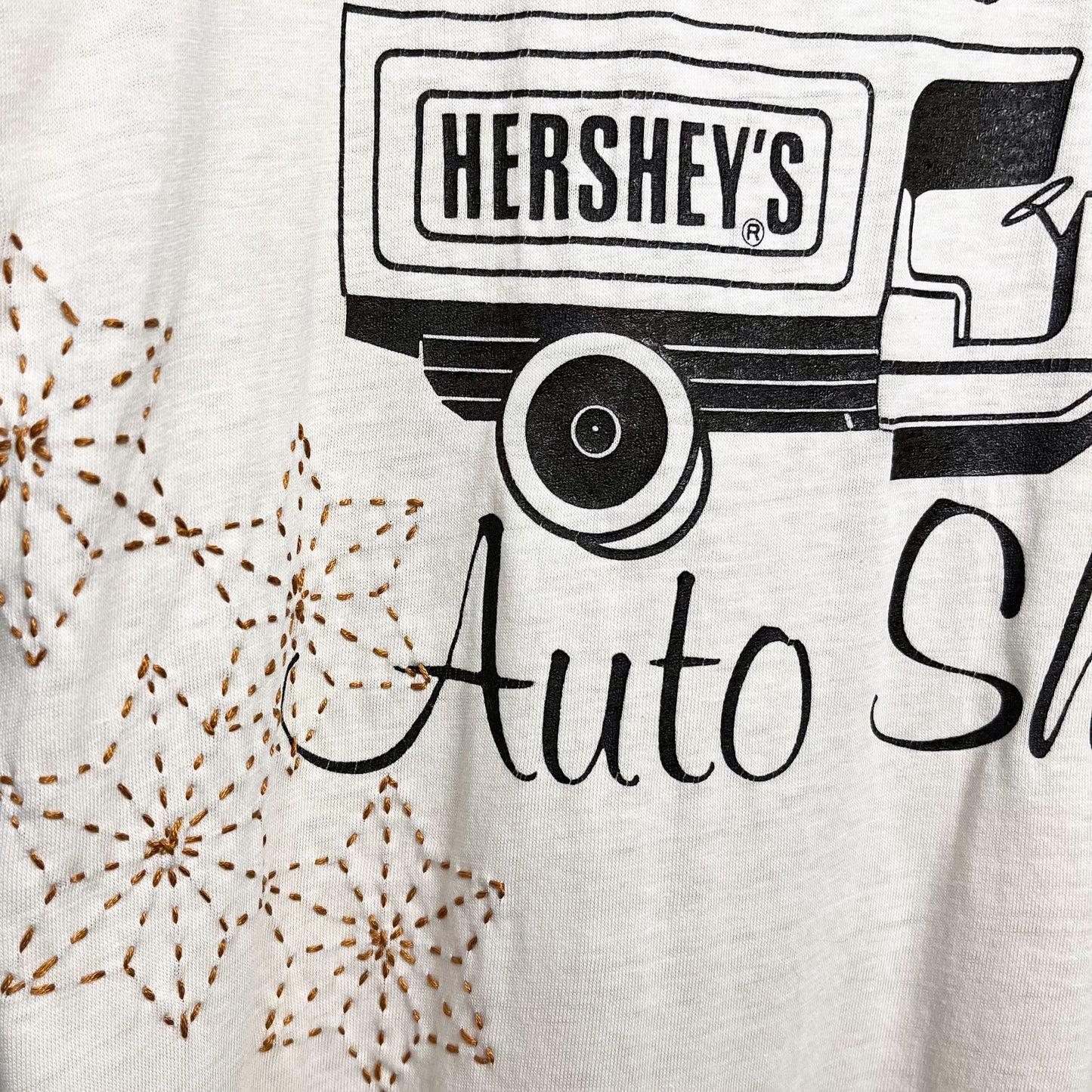 Close up view of a cream colored T-shirt, the t-shirt has a graphic print of a truck that says Hershey's on the back, and the word Auto  below the truck, next to the word Auto there are hand embroidered sashiko stars in mustard brown