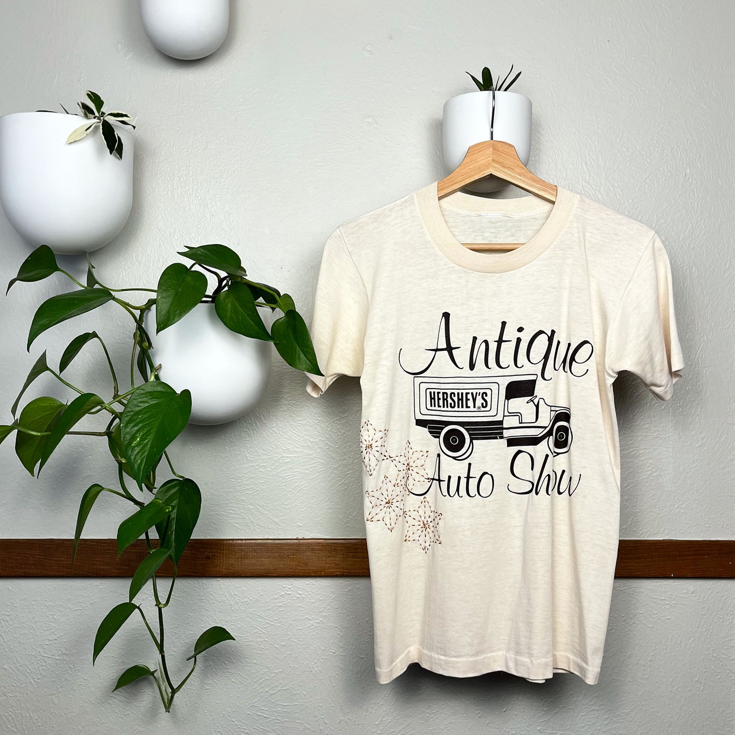 Cream colored T-shirt hanging on a hanger next to a pothos plant on a wall, the t-shirt has a graphic print of a truck that says Hershey's on the back, and the words Antique Auto Show printed around it, on the bottom side of the tee there are hand embroidered sashiko stars in mustard brown