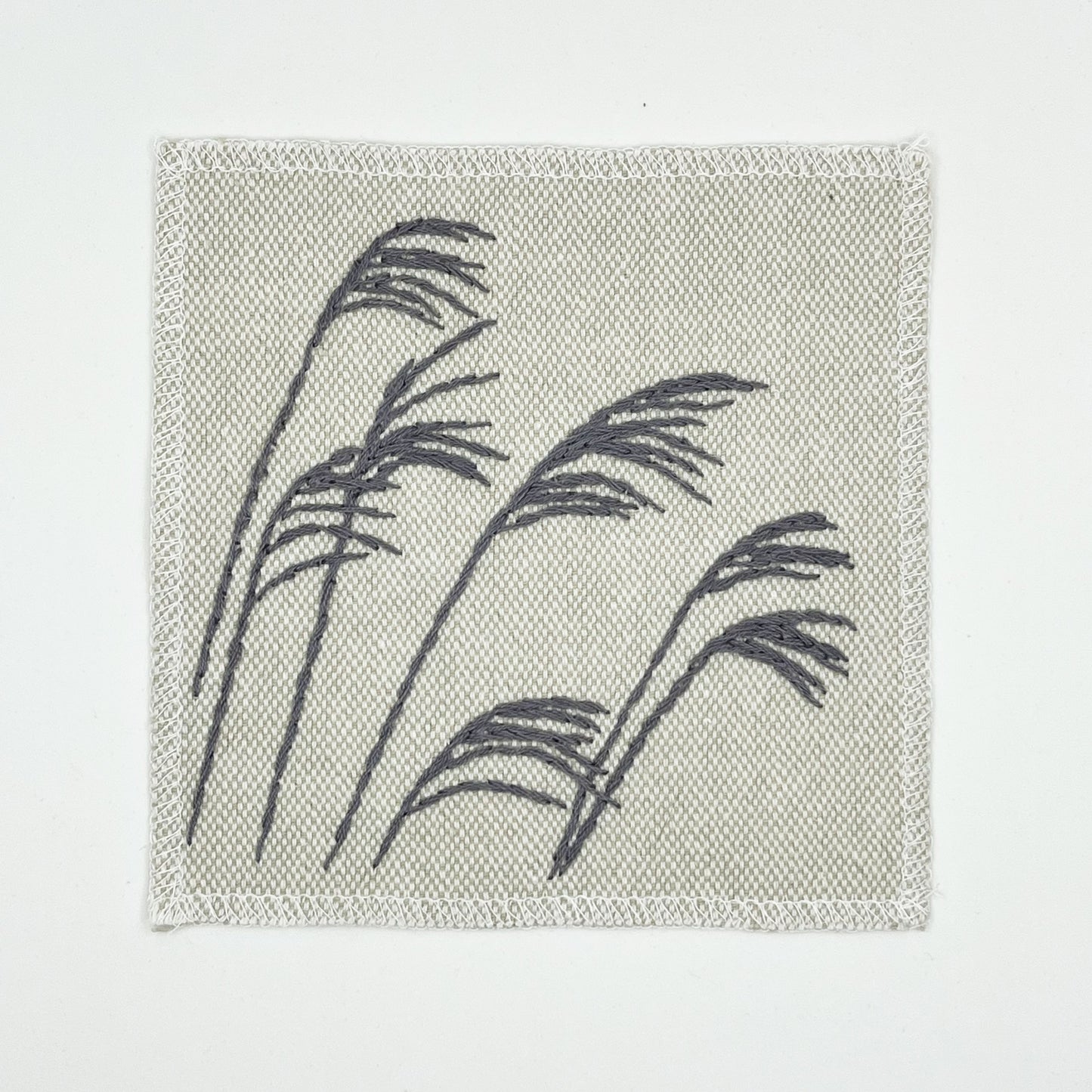 a cream colored square patch with stalks of wild grass stitched in grey thread with a stem stitch, on a white background