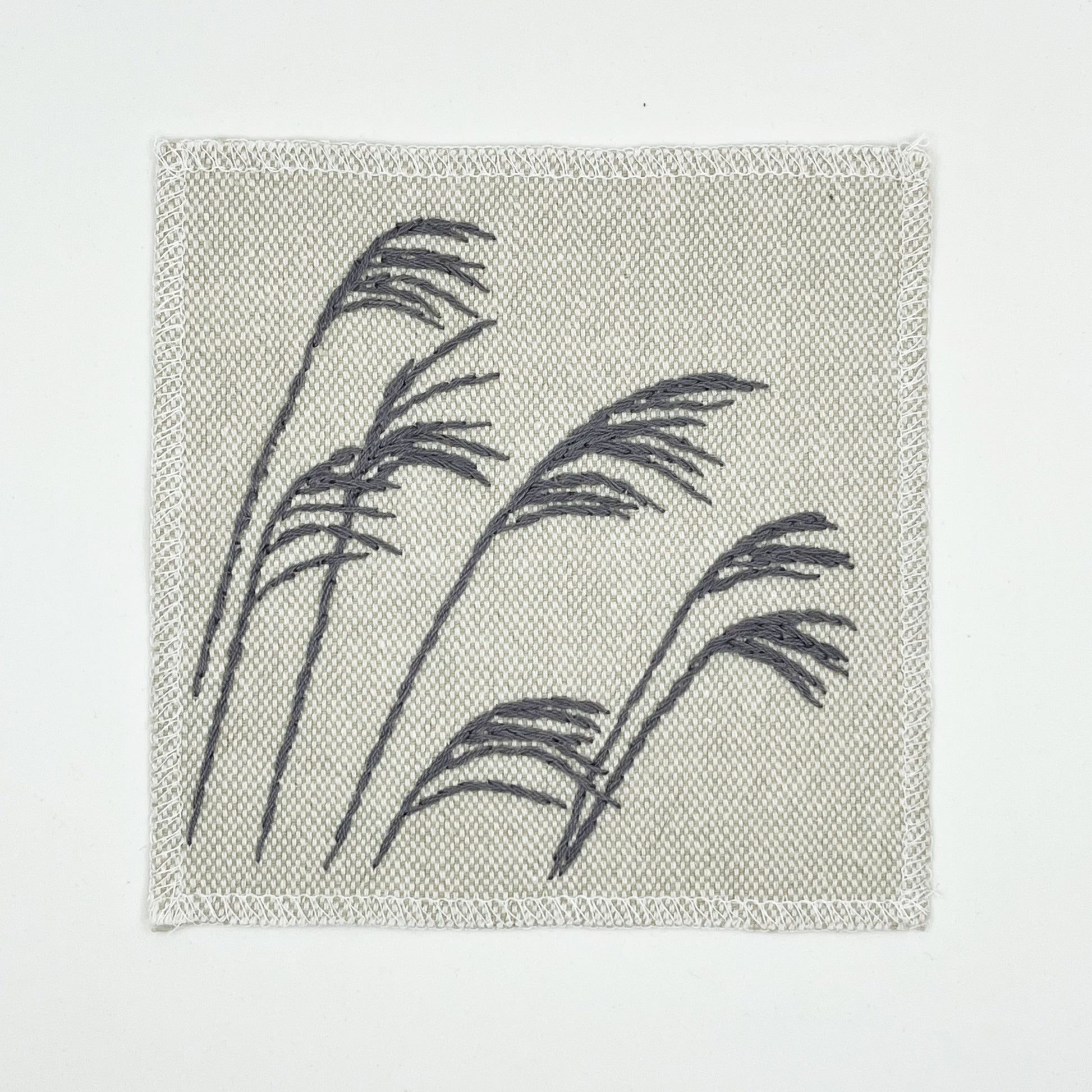 a cream colored square patch with stalks of wild grass stitched in grey thread with a stem stitch, on a white background
