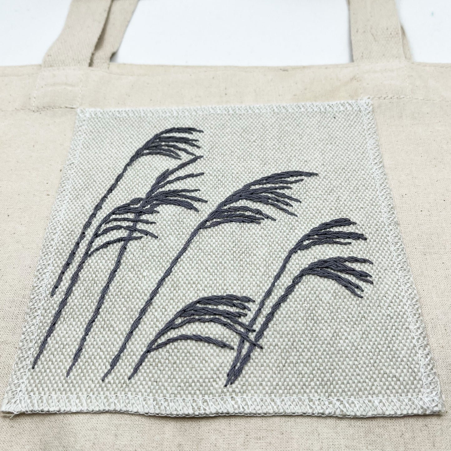 Cream colored square patch with stalks of wild grass stitched in grey thread with a stem stitch, on cream colored tote bag on a white background