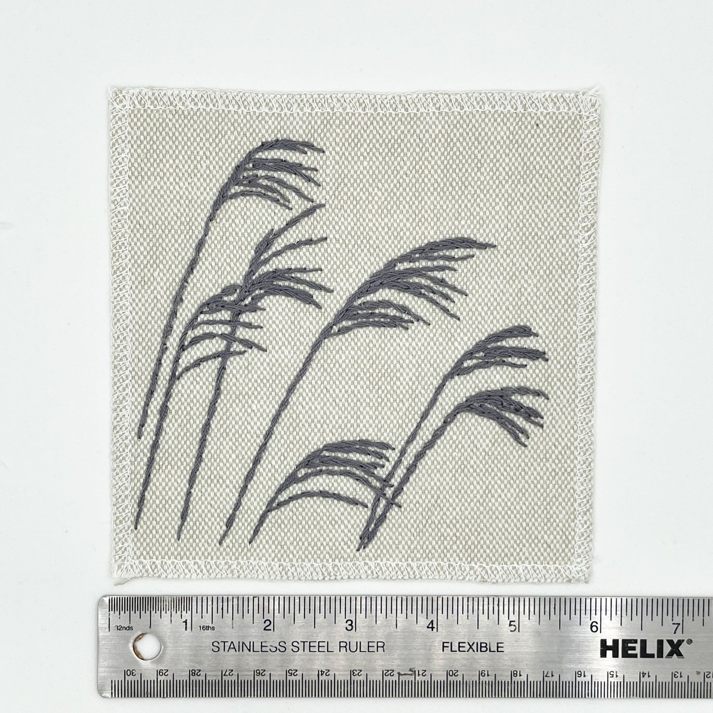 a cream colored square patch with stalks of wild grass stitched in grey thread with a stem stitch, placed next to a metal ruler to show a width of 6 inches, on a white background