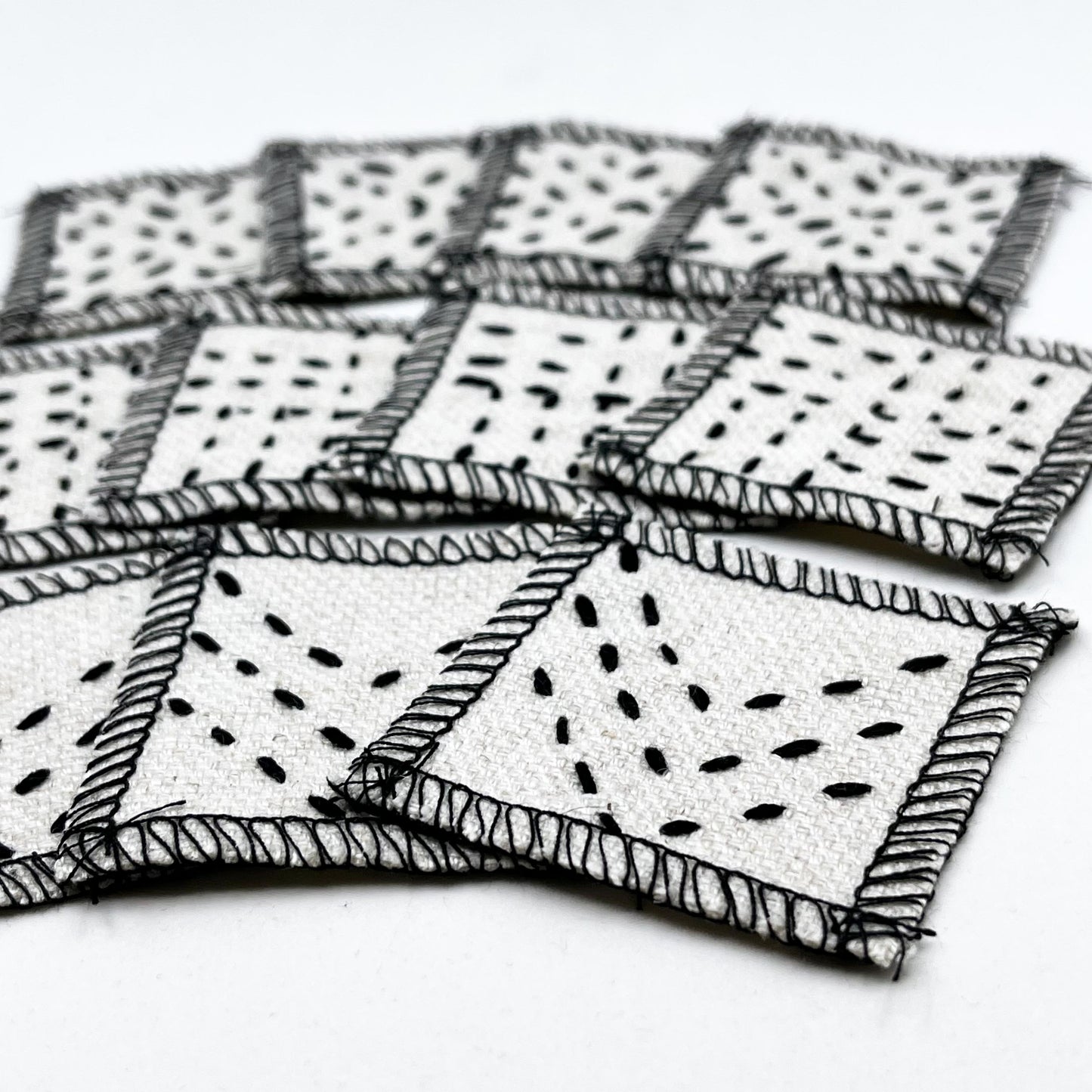 group of eleven square natural colored patches with stitching in black of different patterns, next to a stamp for size comparison