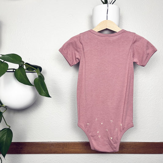 Mauve baby one piece on a hanger, hanging on a wall decorated with plants, with small peach triangle stitches scattered over the bum