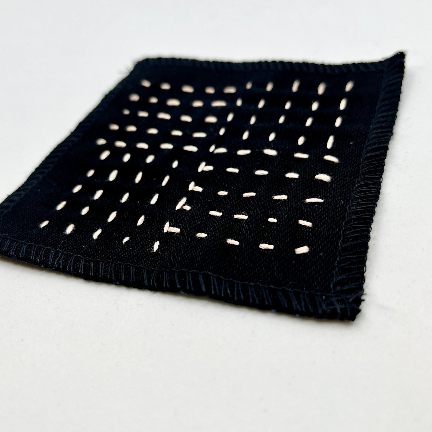 close up angled view of a square black denim patch hand stitched in peach with sashiko style running stitches in the pattern of a basket weave on a white background