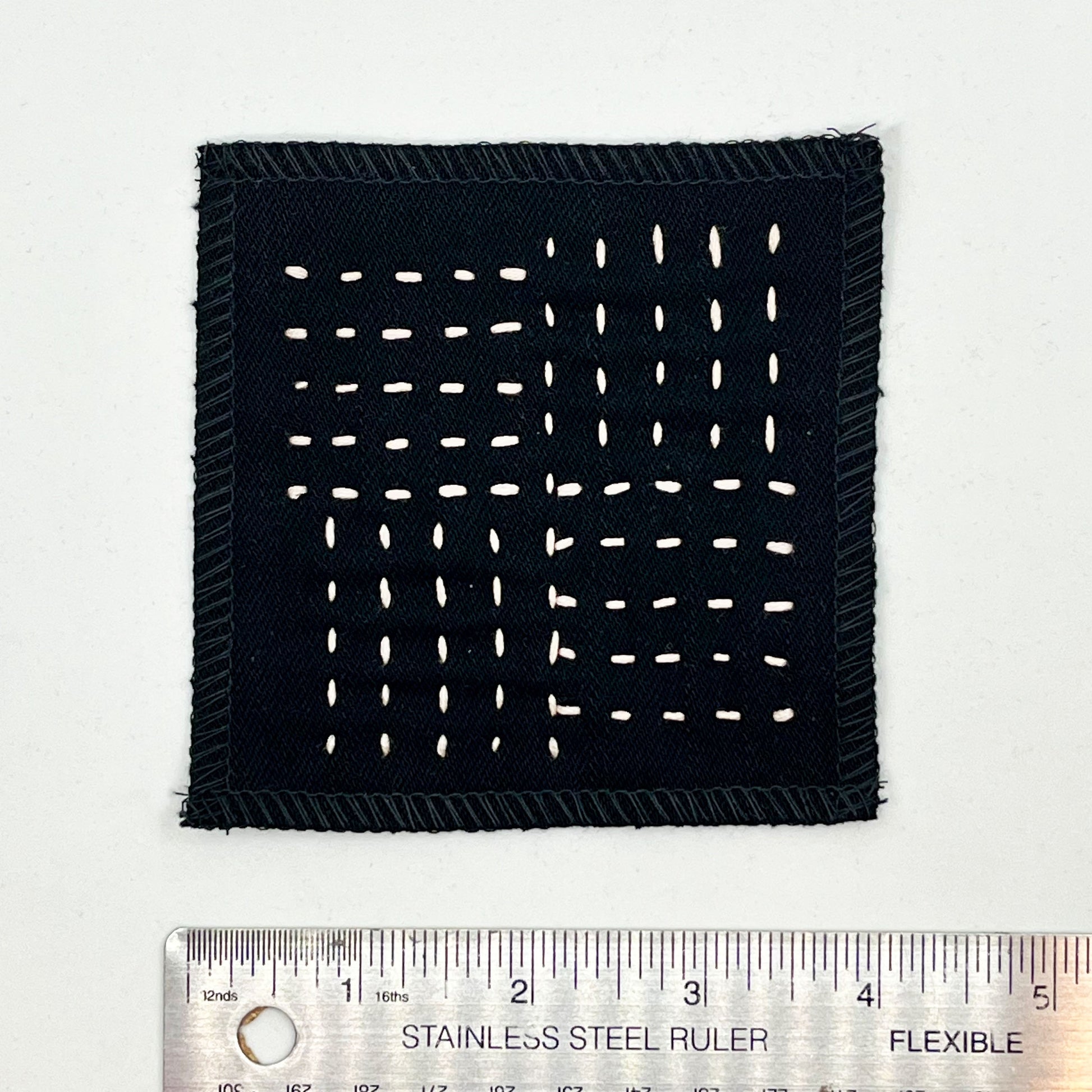a square black denim patch hand stitched in peach with sashiko style running stitches in the pattern of a basket weave placed next to a ruler to show a width of four inches