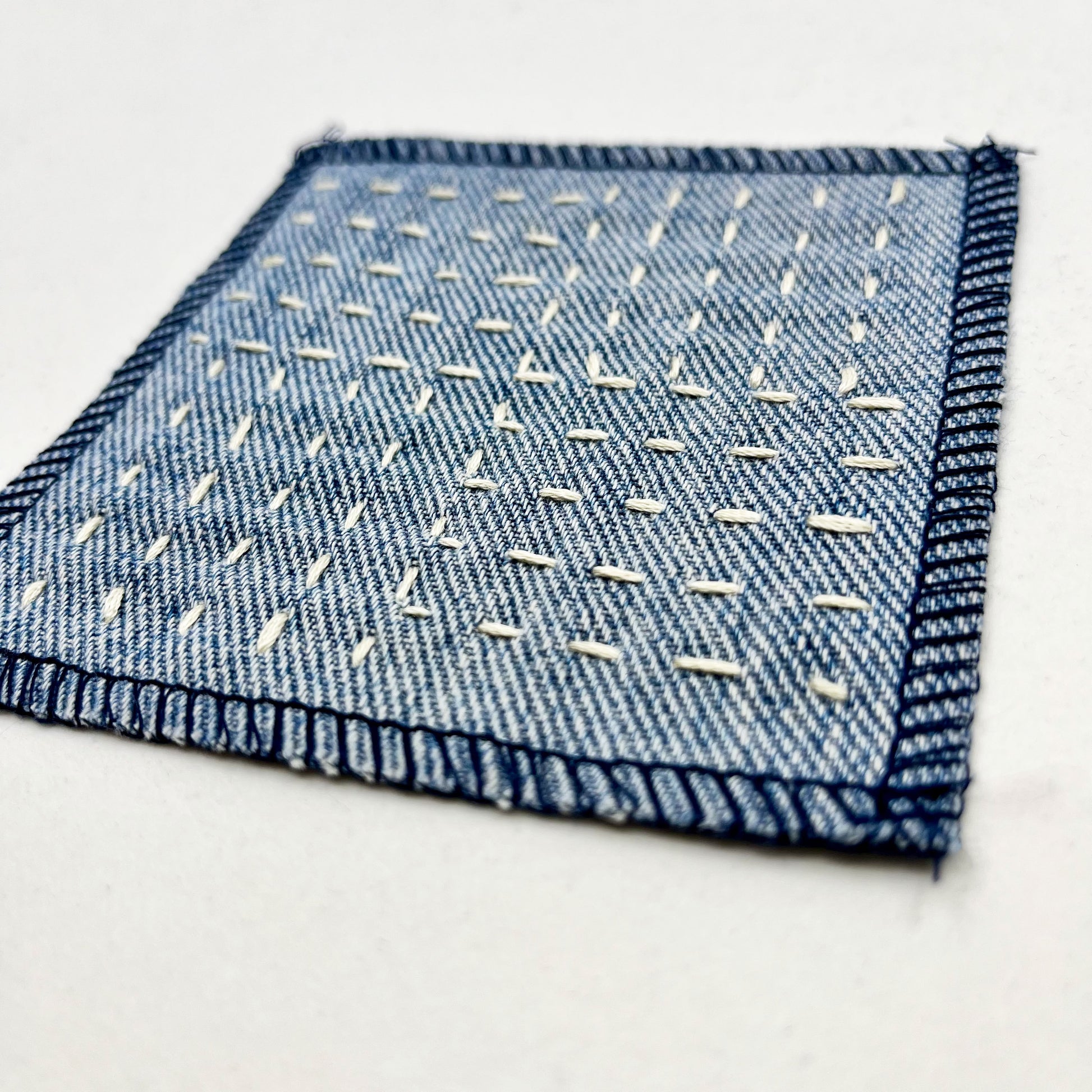 close up angled view of a square denim patch hand stitched in ivory with sashiko style running stitches in the pattern of a basket weave  on a white background