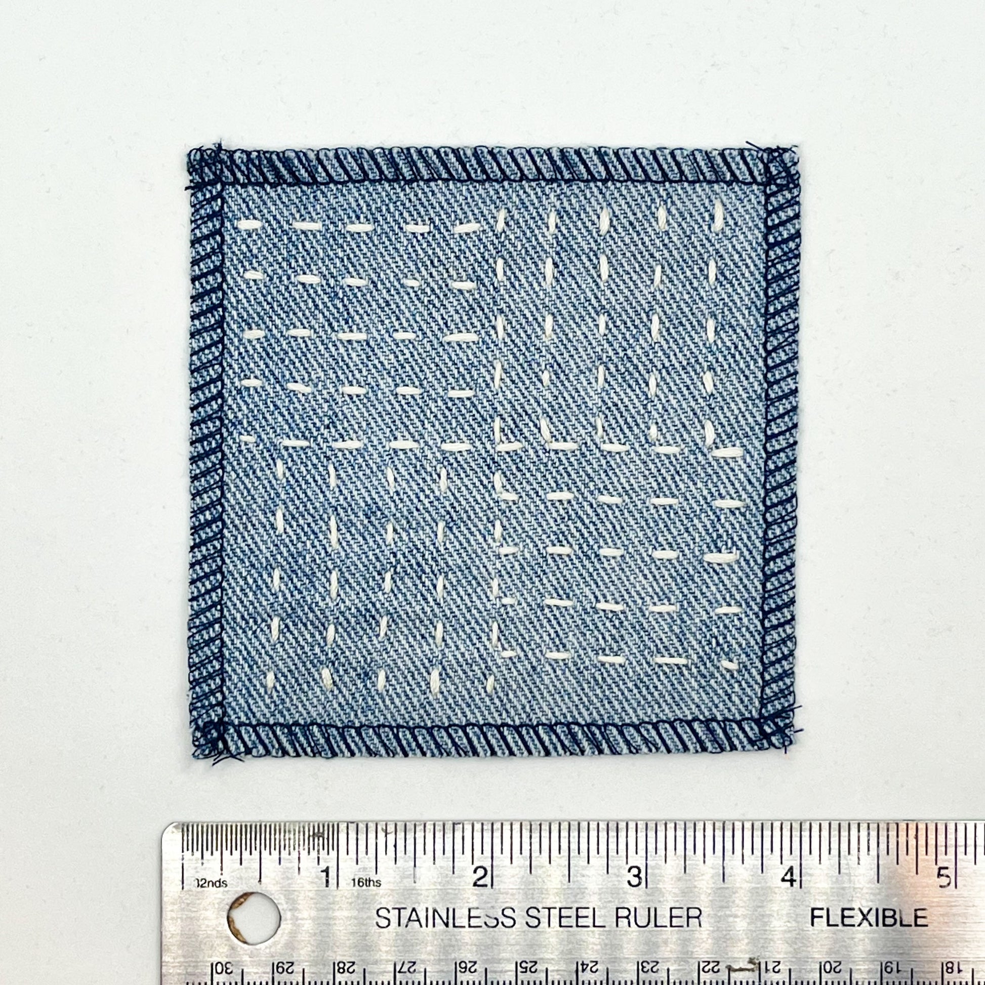 a square denim patch hand stitched in ivory with sashiko style running stitches in the pattern of a basket weave placed next to a ruler to show a width of four inches