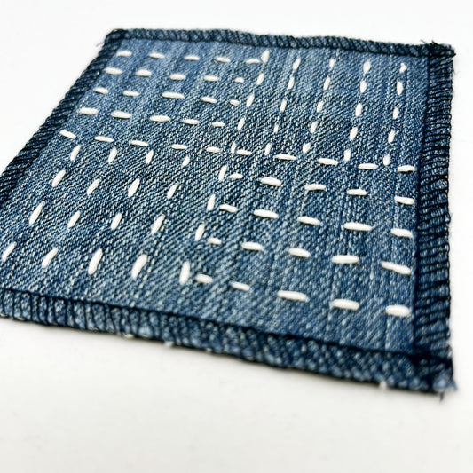 close up angled view of a square denim patch hand stitched in ivory with sashiko style running stitches in the pattern of a basket weave on a white background