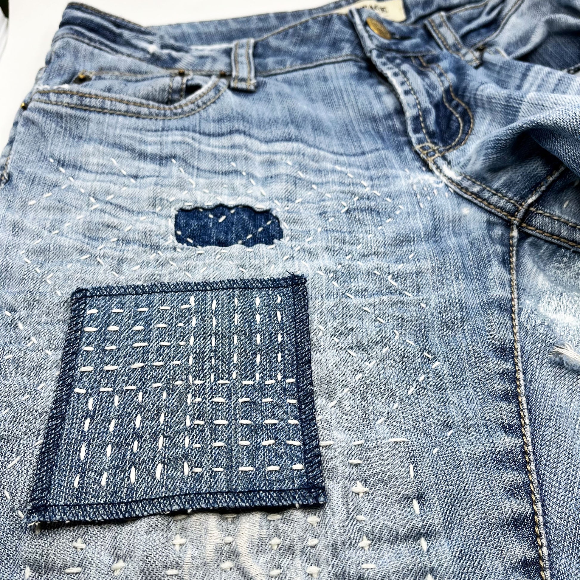 a square denim patch hand stitched in ivory with sashiko style running stitches in the pattern of a basket weave on a pair of heavily mended jeans