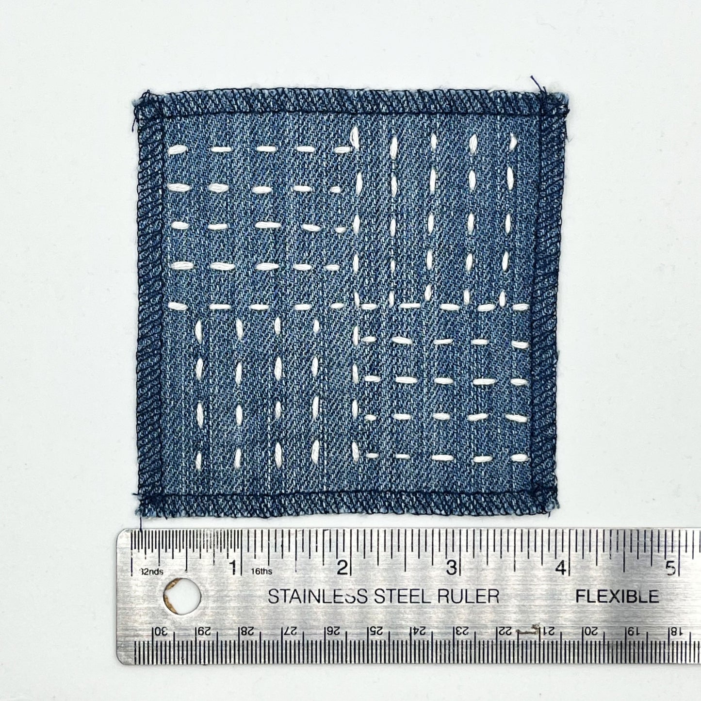 a square denim patch hand stitched in ivory with sashiko style running stitches in the pattern of a basket weave placed next to a metal ruler to show a width of four inches