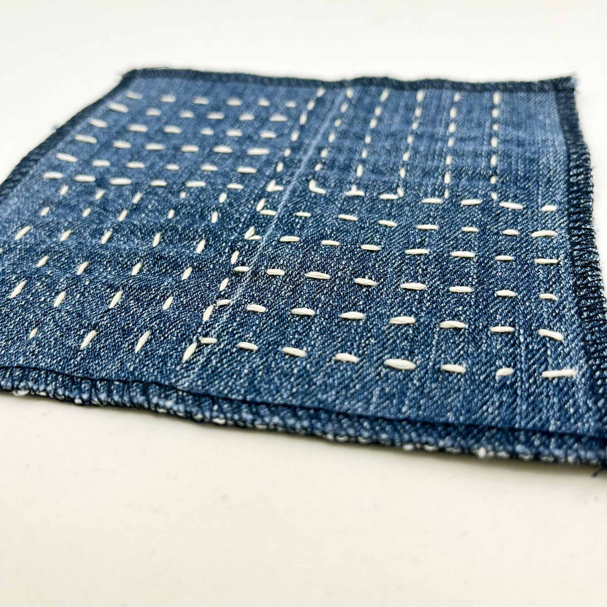 close up angled view of a square denim patch hand stitched in ivory with sashiko style running stitches in the pattern of a basket weave on a white background