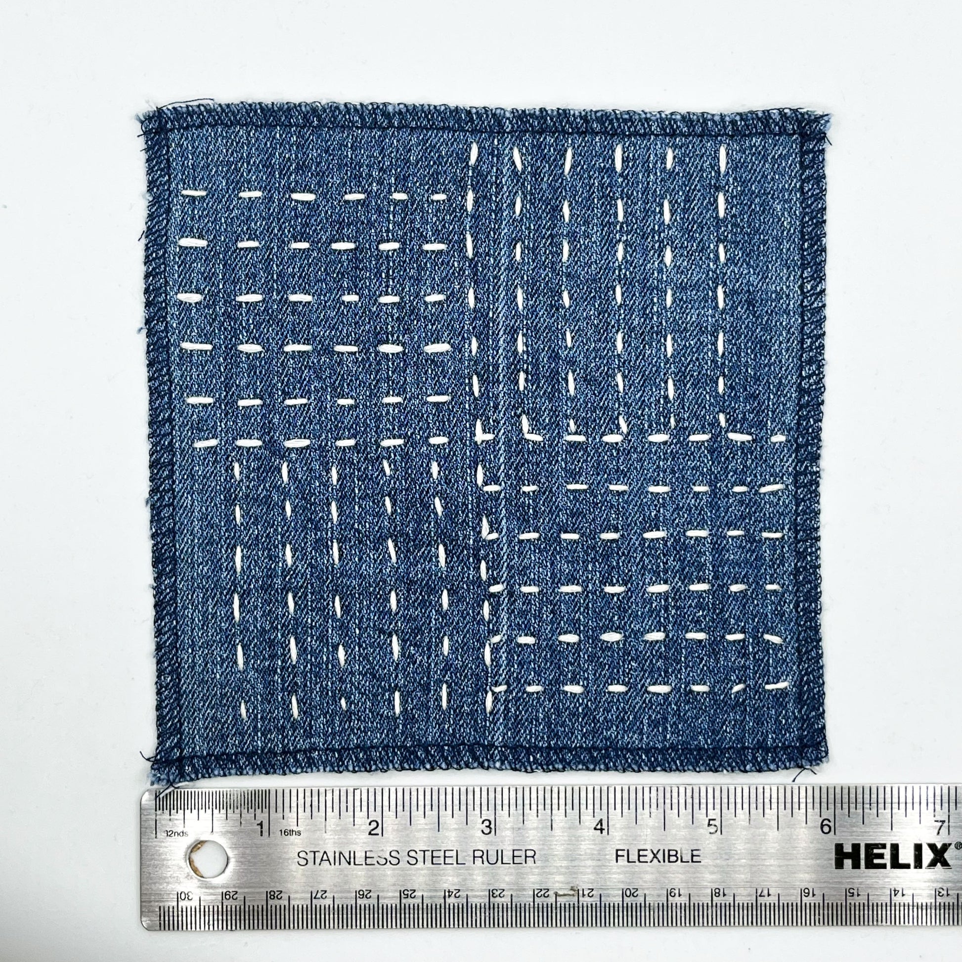 a square denim patch hand stitched in ivory with sashiko style running stitches in the pattern of a basket weave next to a metal ruler to show a width of six inches
